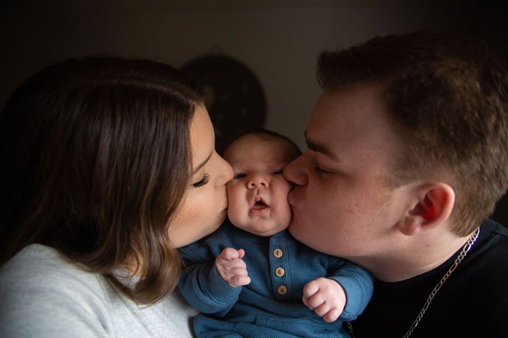 Parents share loving moment kissing their baby in denim clothing during intimate family portrait session.