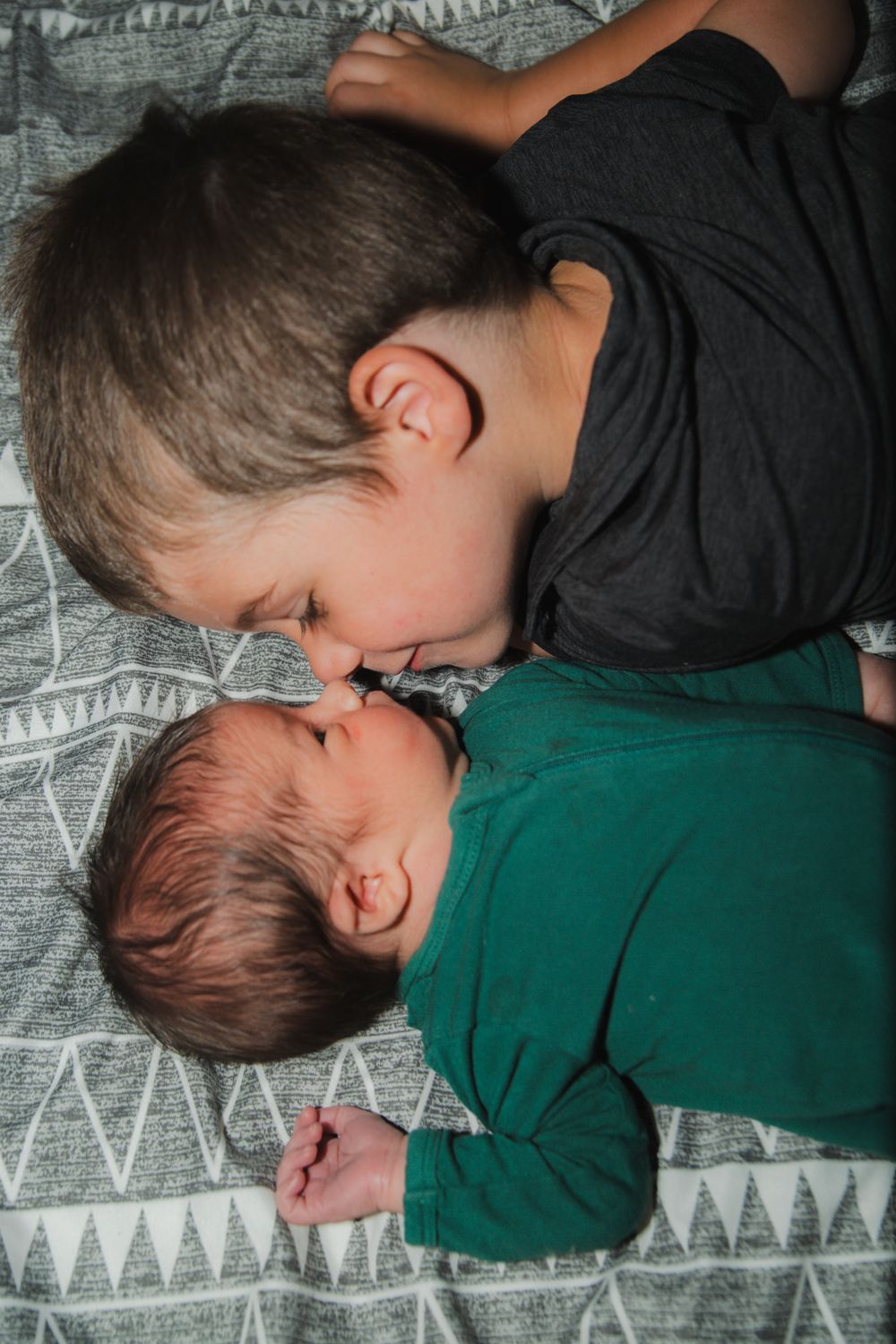 Two children in black and green shirts lying close together on a geometric patterned blanket.
