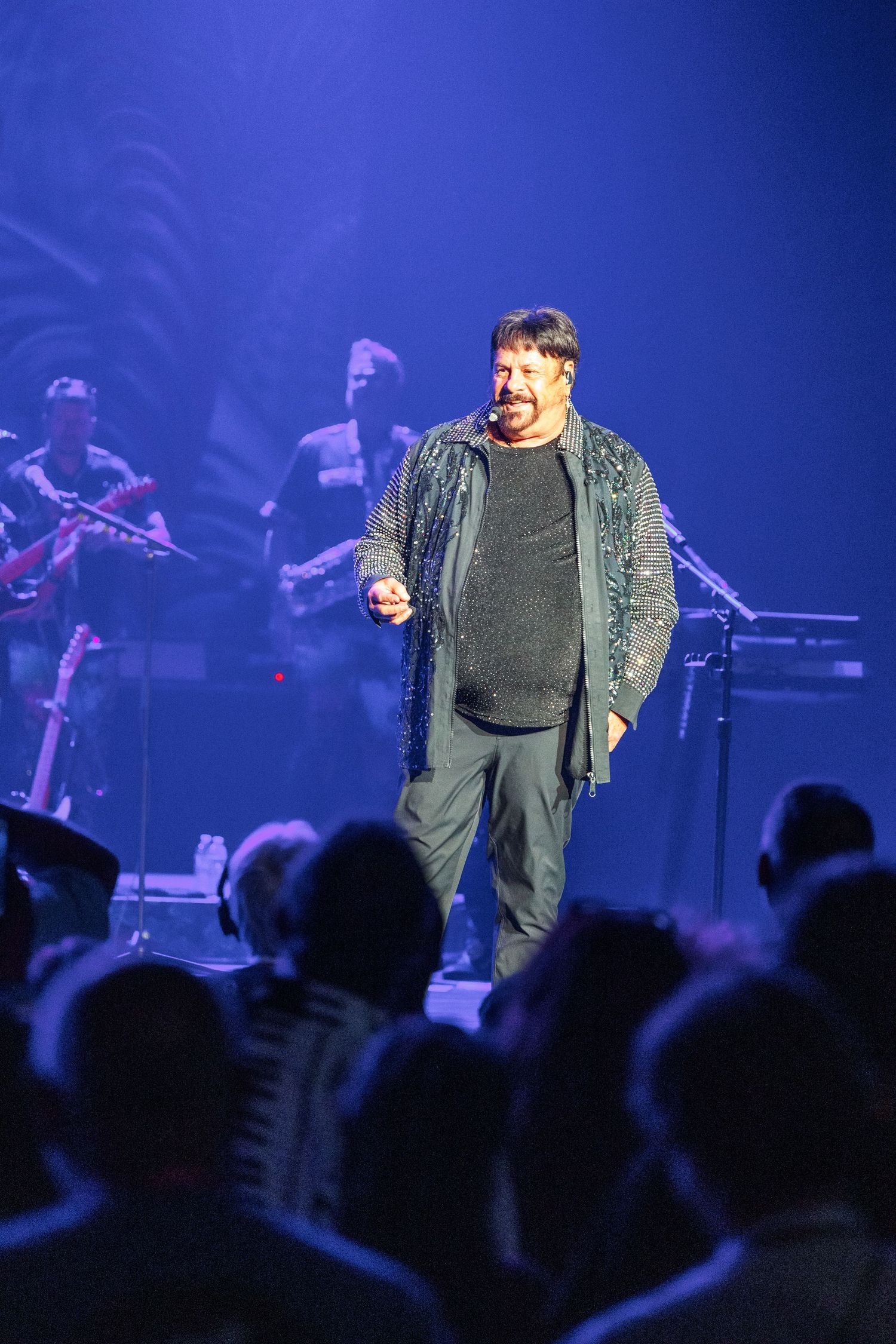 A performer in a gray sweater and jacket stands on stage singing into a microphone with band members and crowd visible.