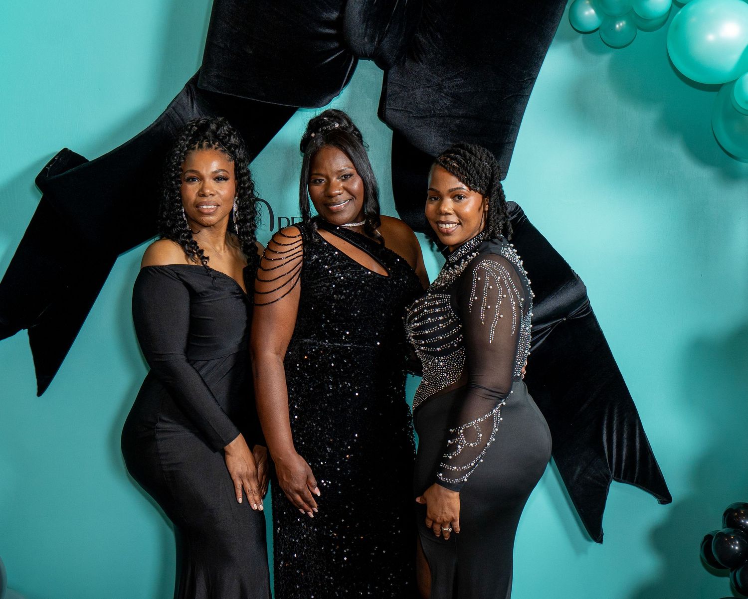Three women in elegant black dresses pose against a turquoise backdrop with dramatic lighting.