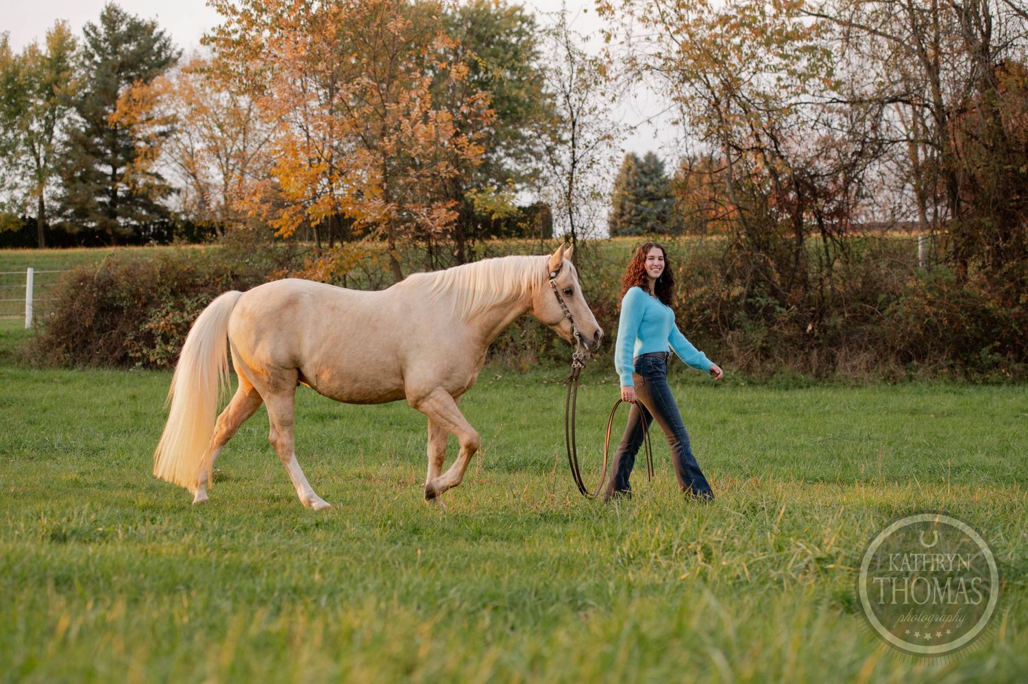 A person leads a cream-colored horse through a grassy field in autumn.