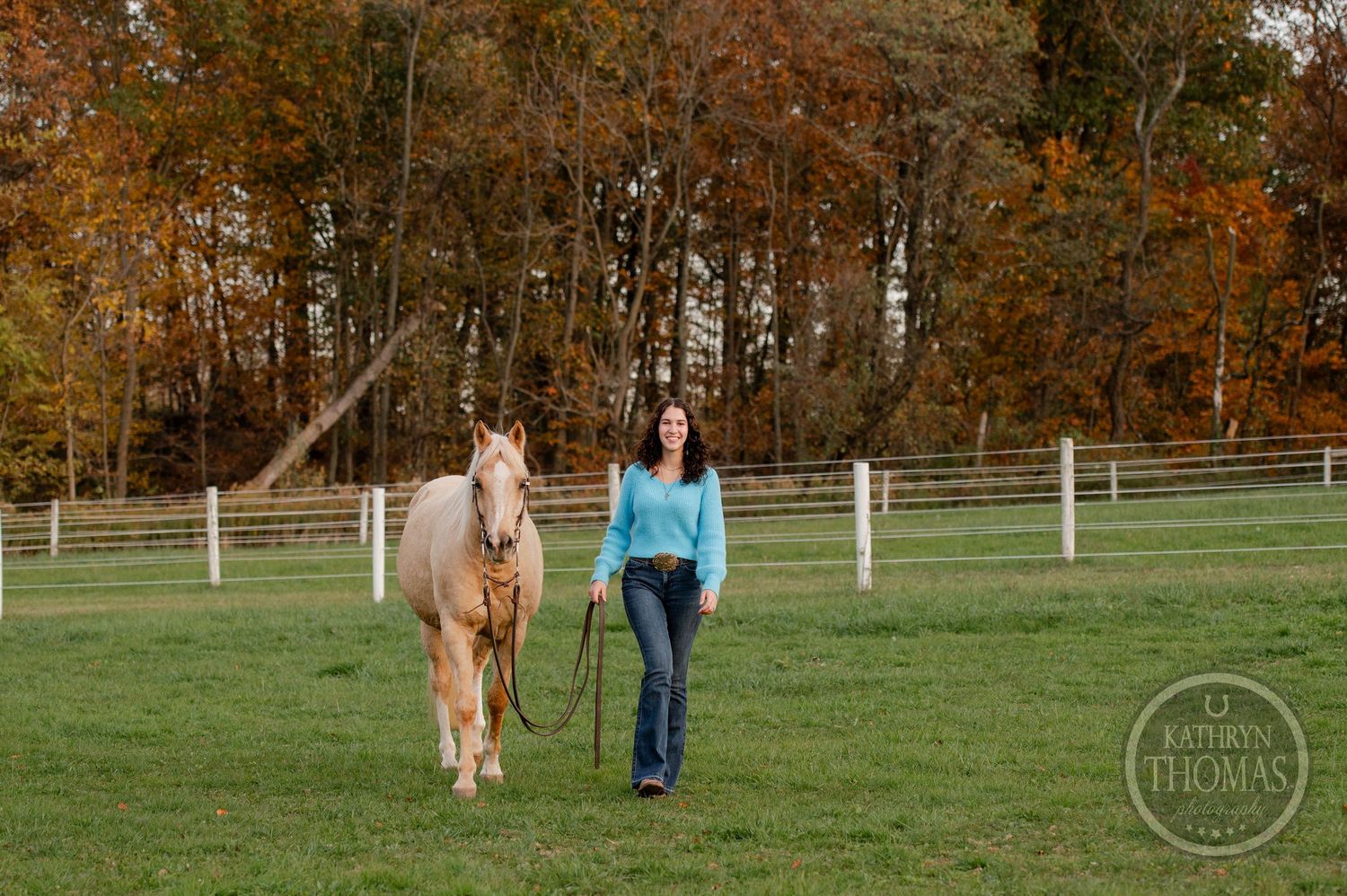 A pale golden horse walks beside someone through a grassy field bordered by fall foliage.