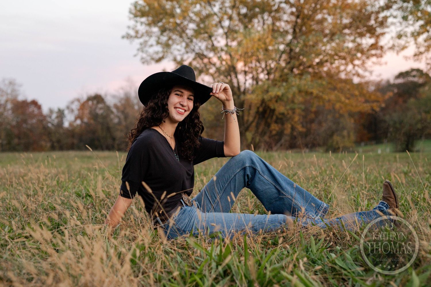 Person in western attire relaxes in autumn field during golden hour photoshoot.