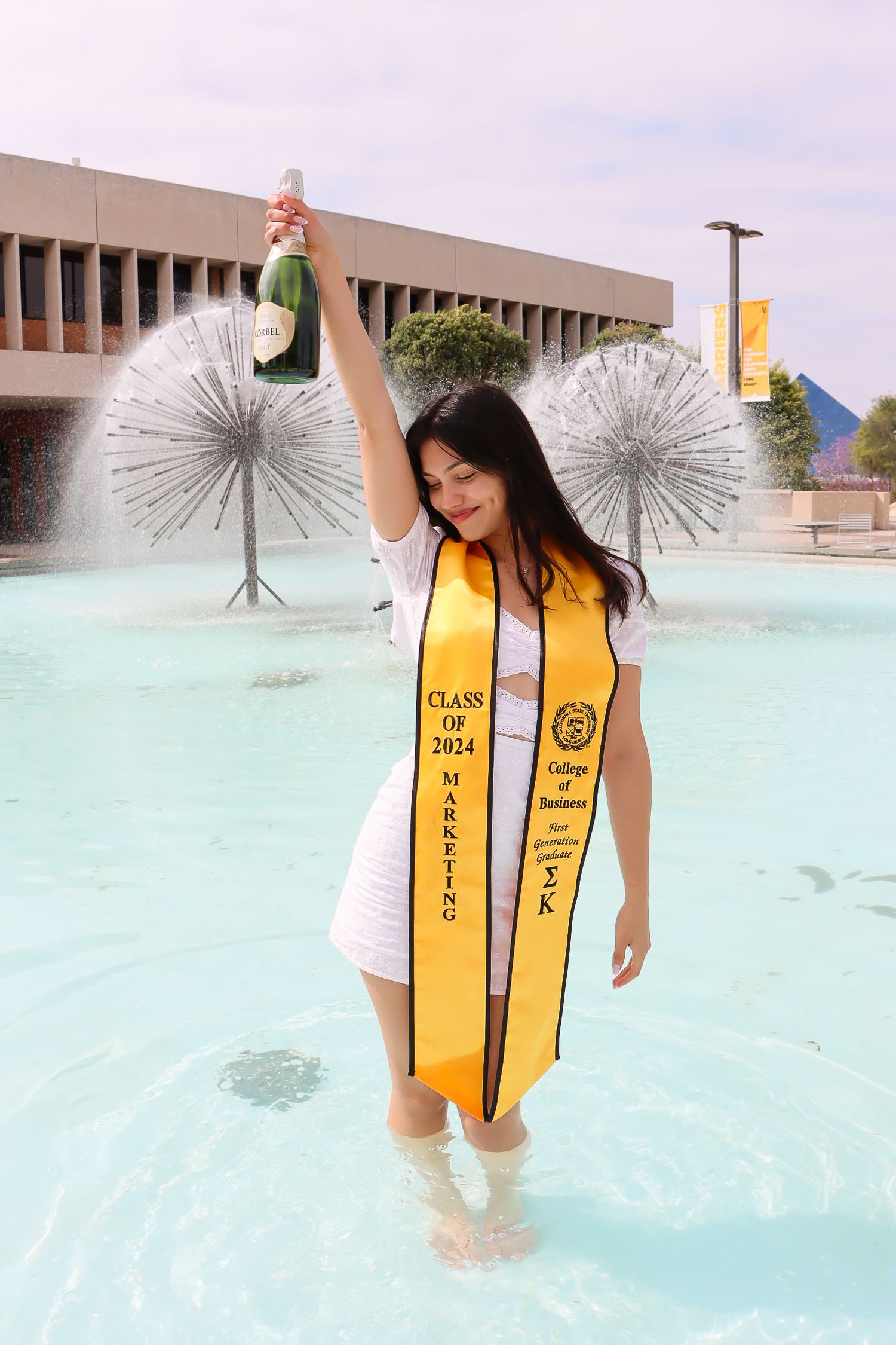 Graduate celebrating in a fountain wearing gold academic stoles at a university campus.