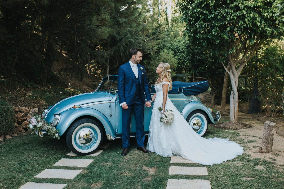 A vintage light blue Volkswagen Beetle parked in a garden setting with a bride and groom standing beside it.