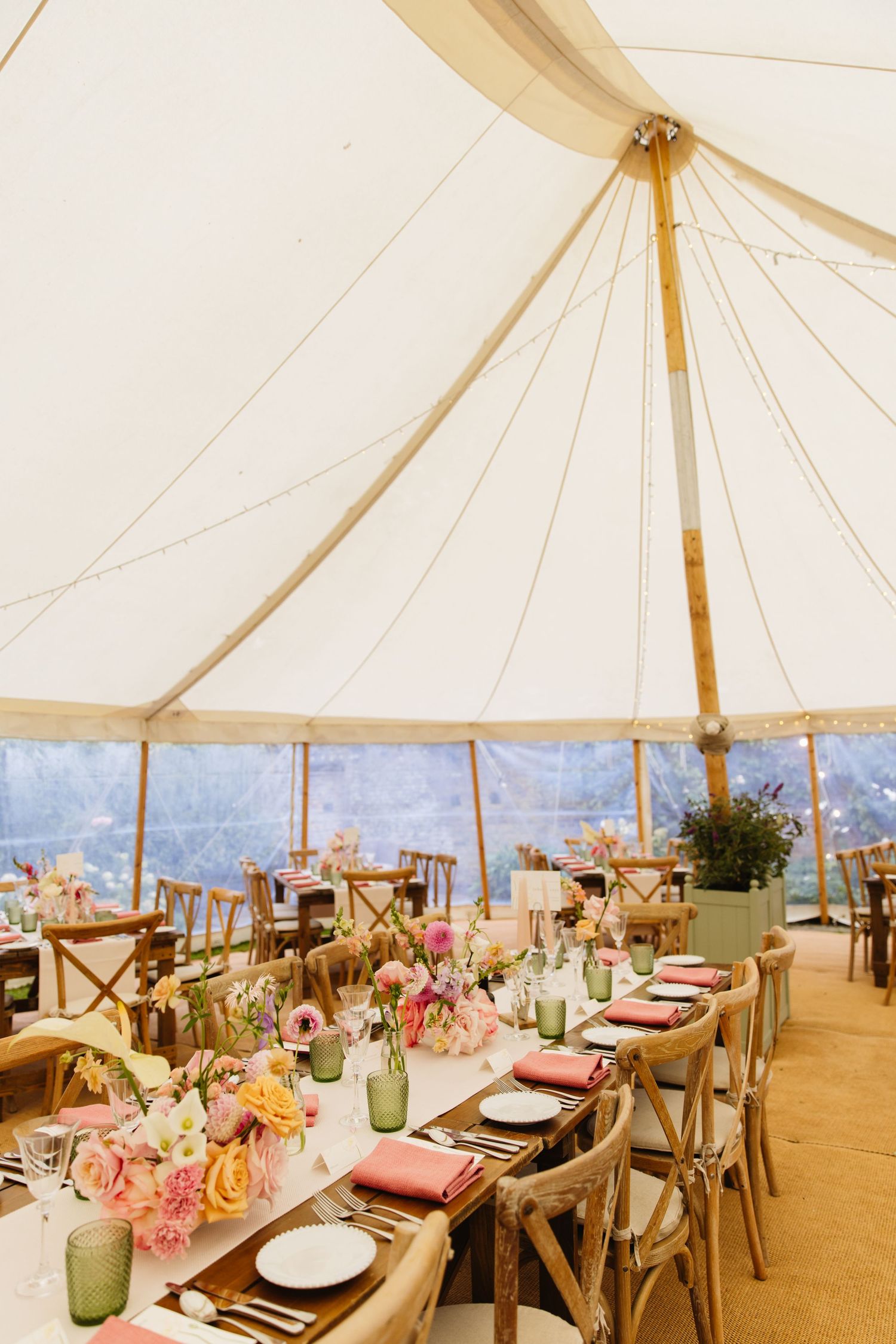 Elegant wedding reception setup inside white tent with rustic wood tables, green glassware and pink floral centerpieces.