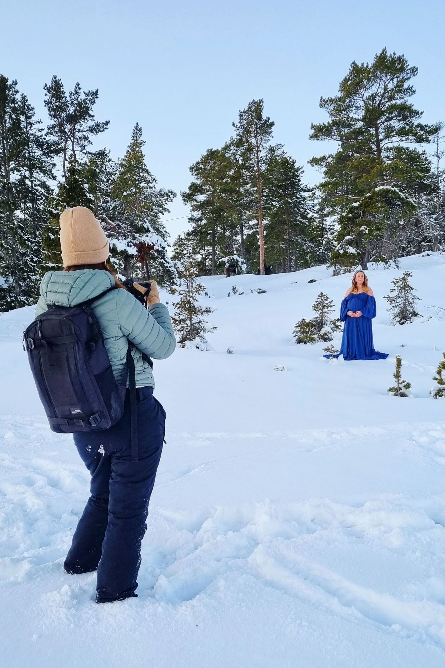 Photographer capturing a woman in a blue dress kneeling in a snowy winter forest landscape.