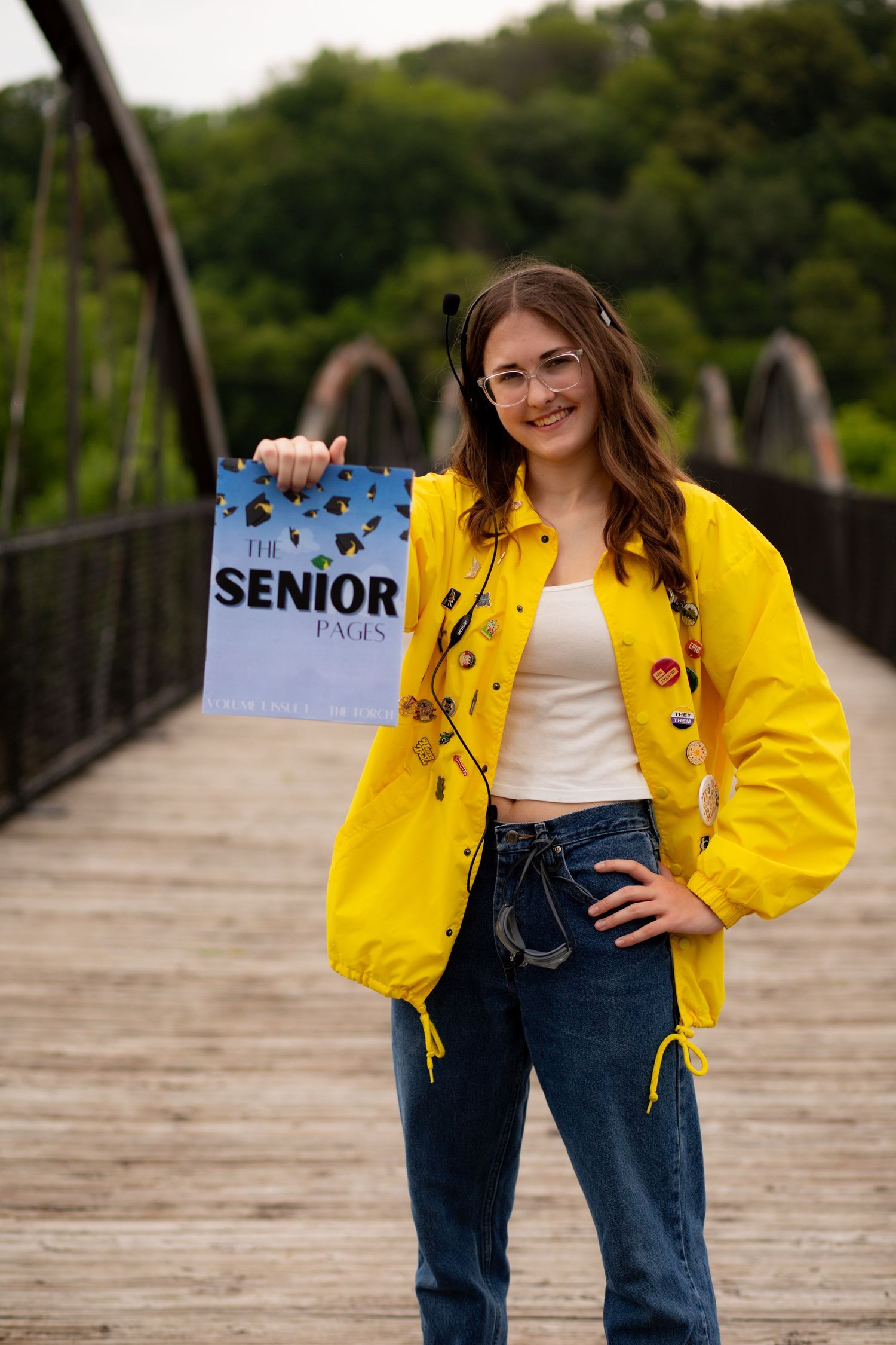 On a wooden bridge, someone in a bright yellow jacket and jeans holds up a senior sign while posing for a photo.