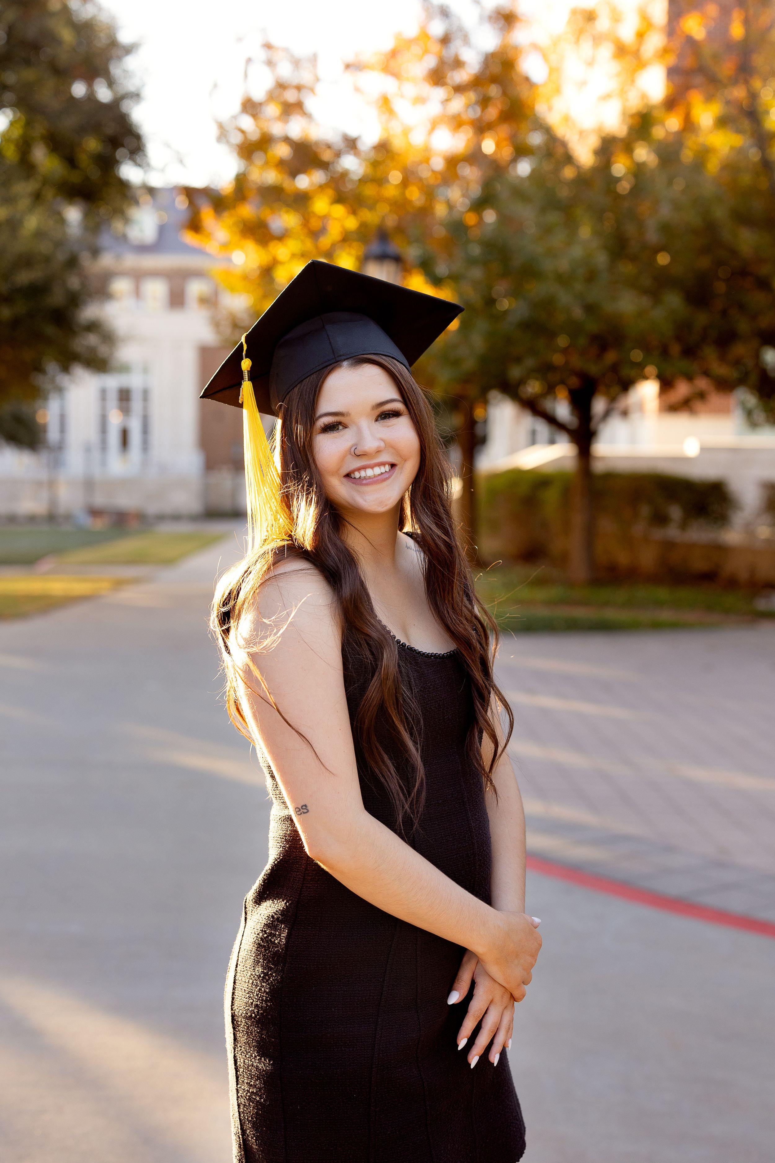 A graduate in black cap and gown poses outdoors during golden hour with autumn trees in the background.