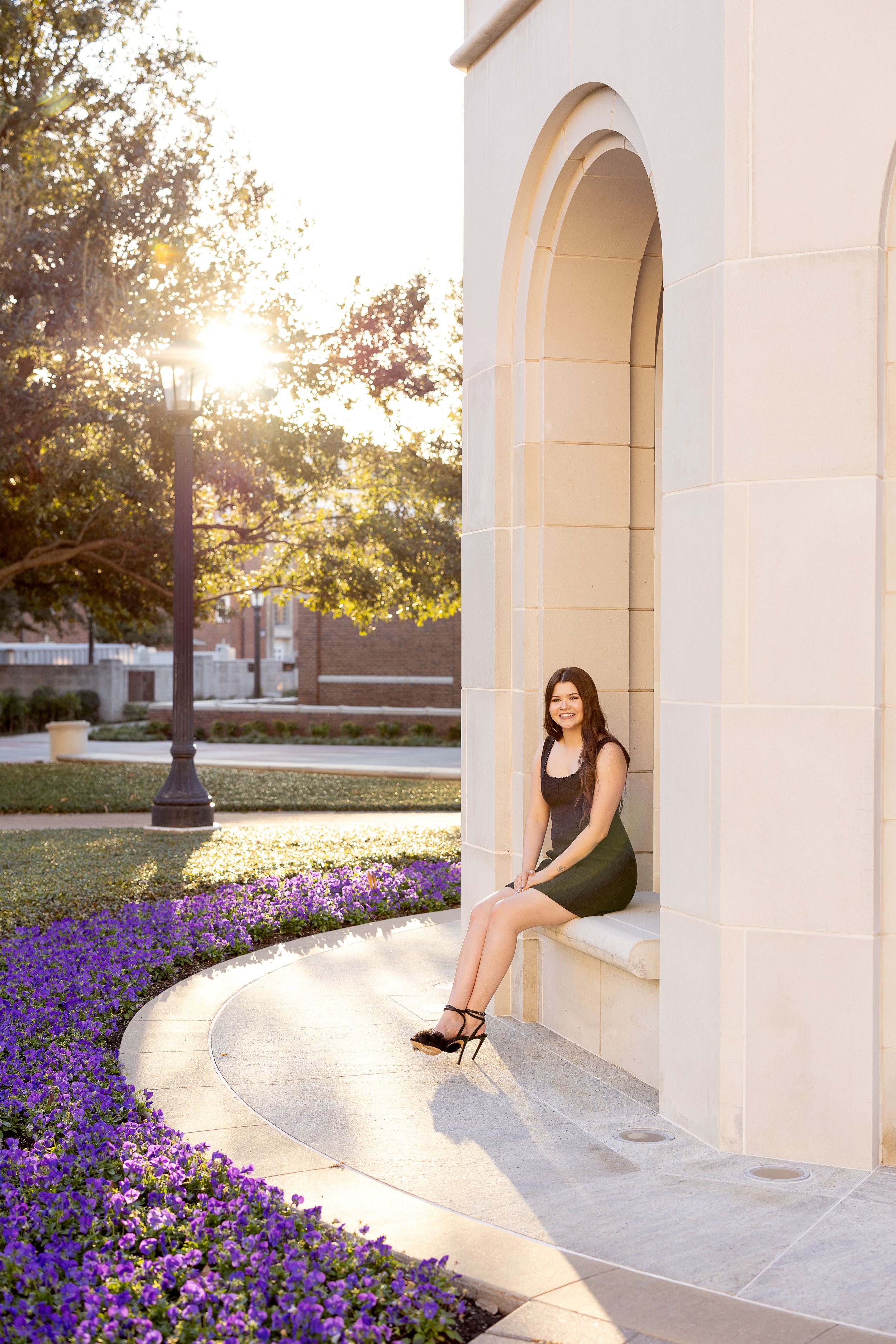 A woman sits in an arched alcove at sunset with purple flowers blooming along a curved garden path.