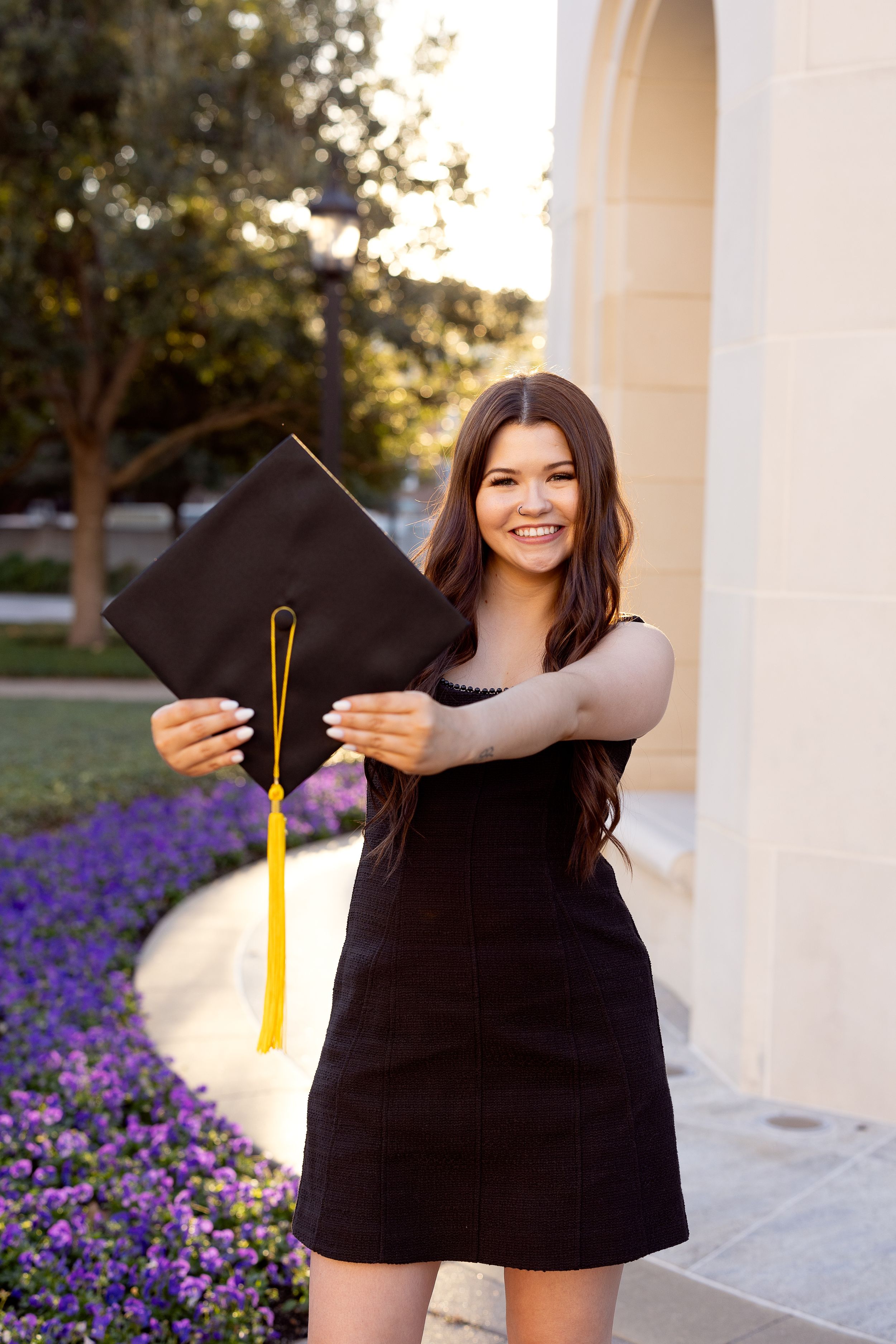 Graduate in black dress holding mortarboard cap with gold tassel poses by purple flowers and white columns outdoors.