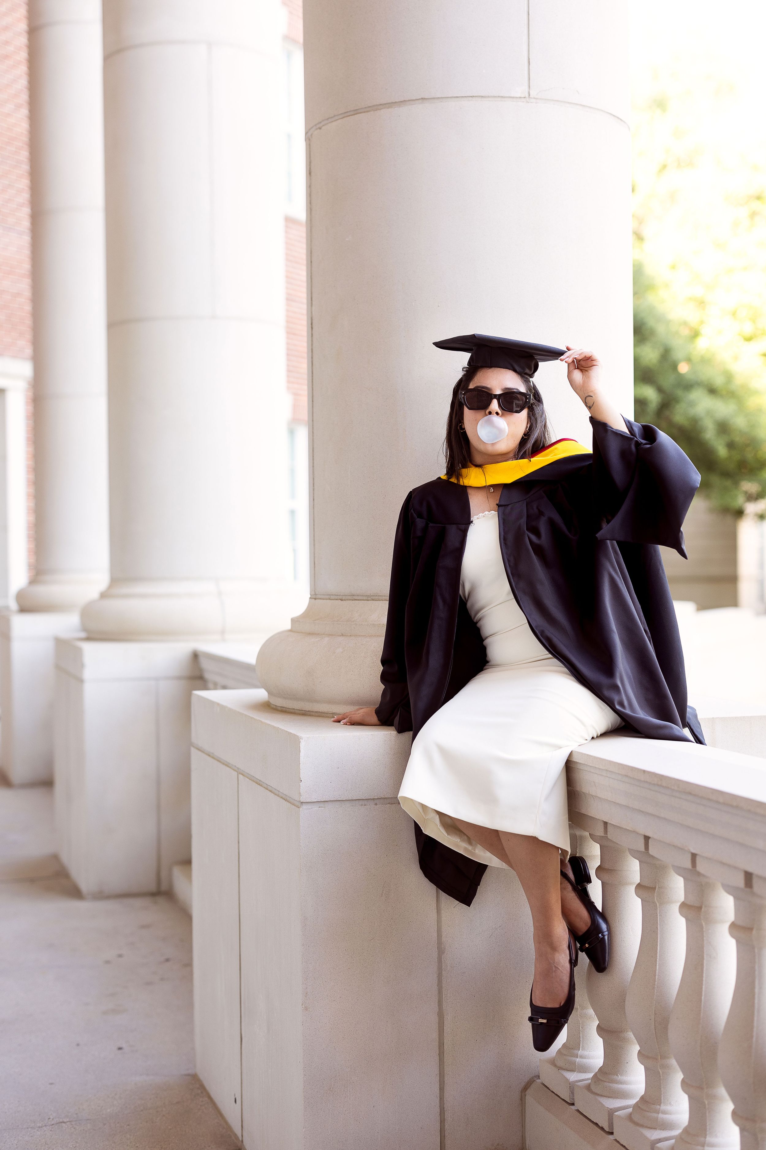 Graduate in cap and gown with yellow stole sitting on white stone balcony railing by classical columns in sunlight.