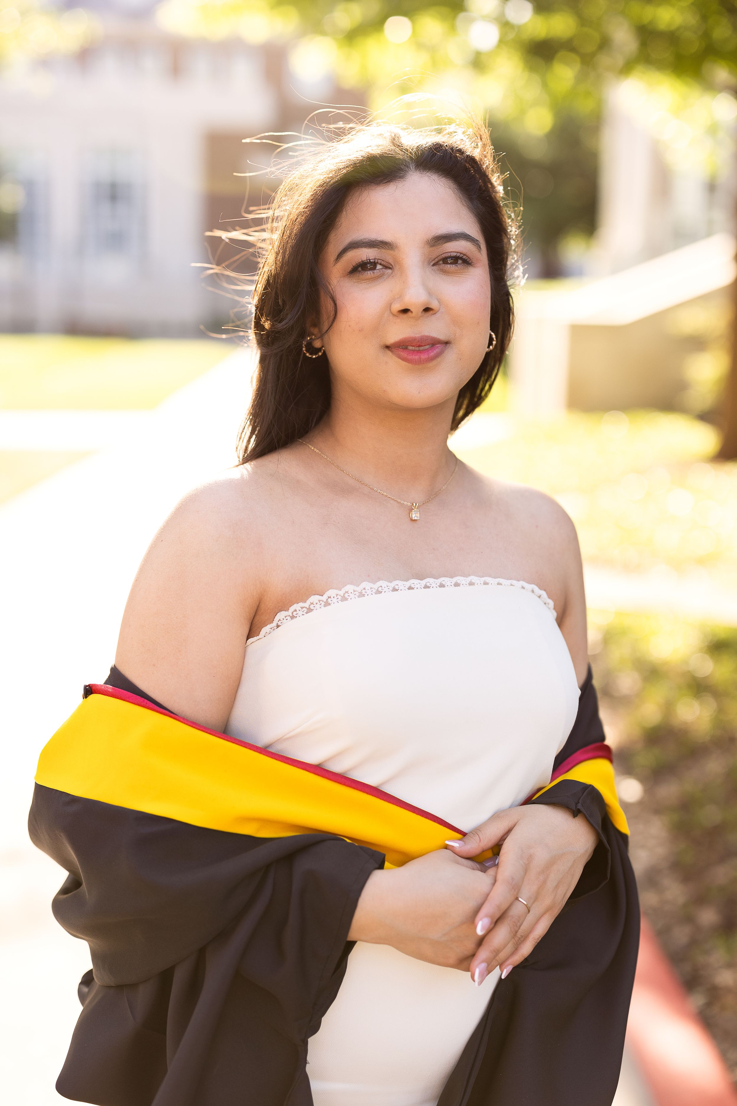 A graduate in a white dress and black and gold graduation gown poses outdoors on a sunny day during commencement.