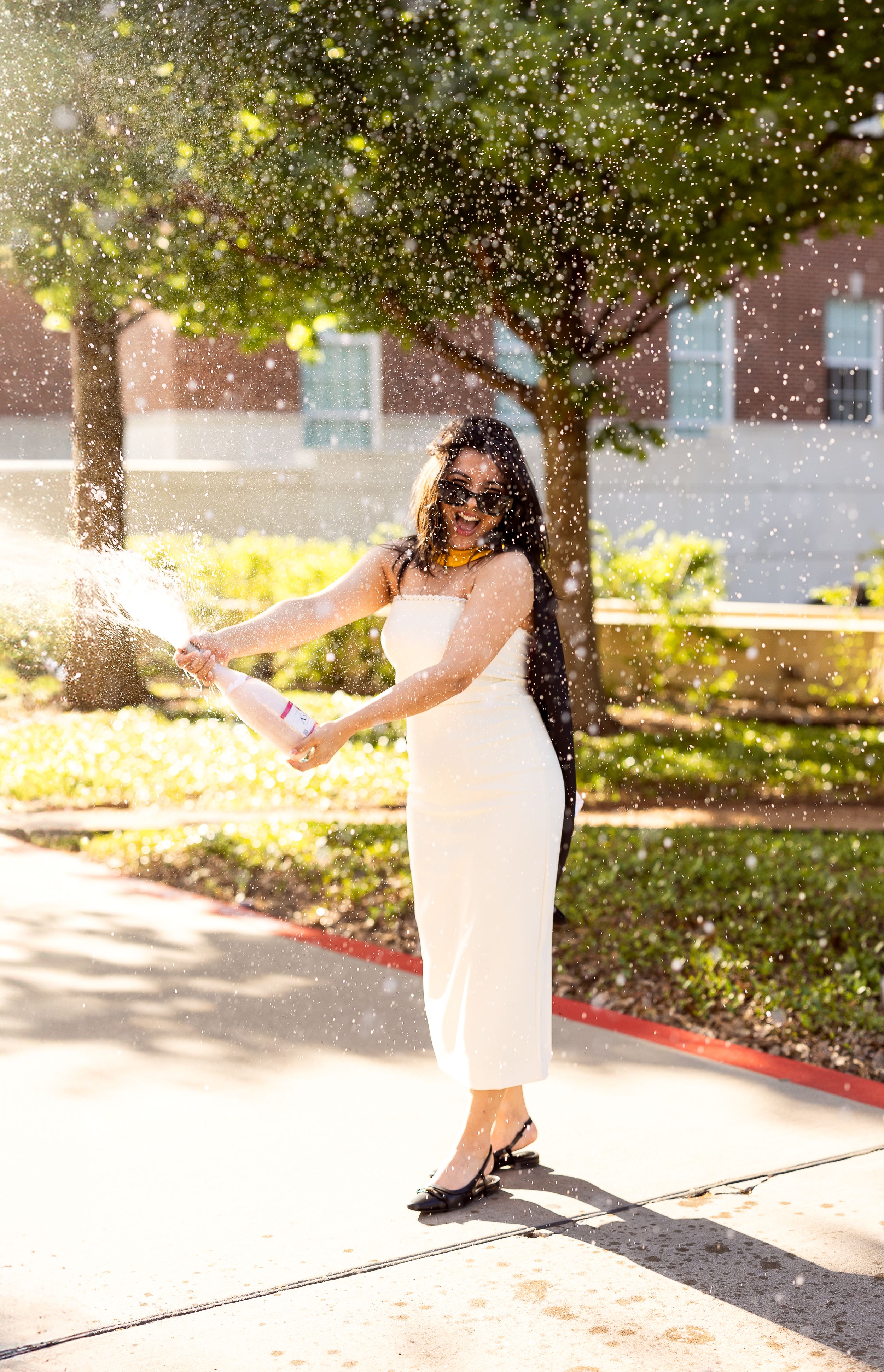 Person in white dress and sunglasses stands outdoors on sunny day with water droplets sparkling in background.