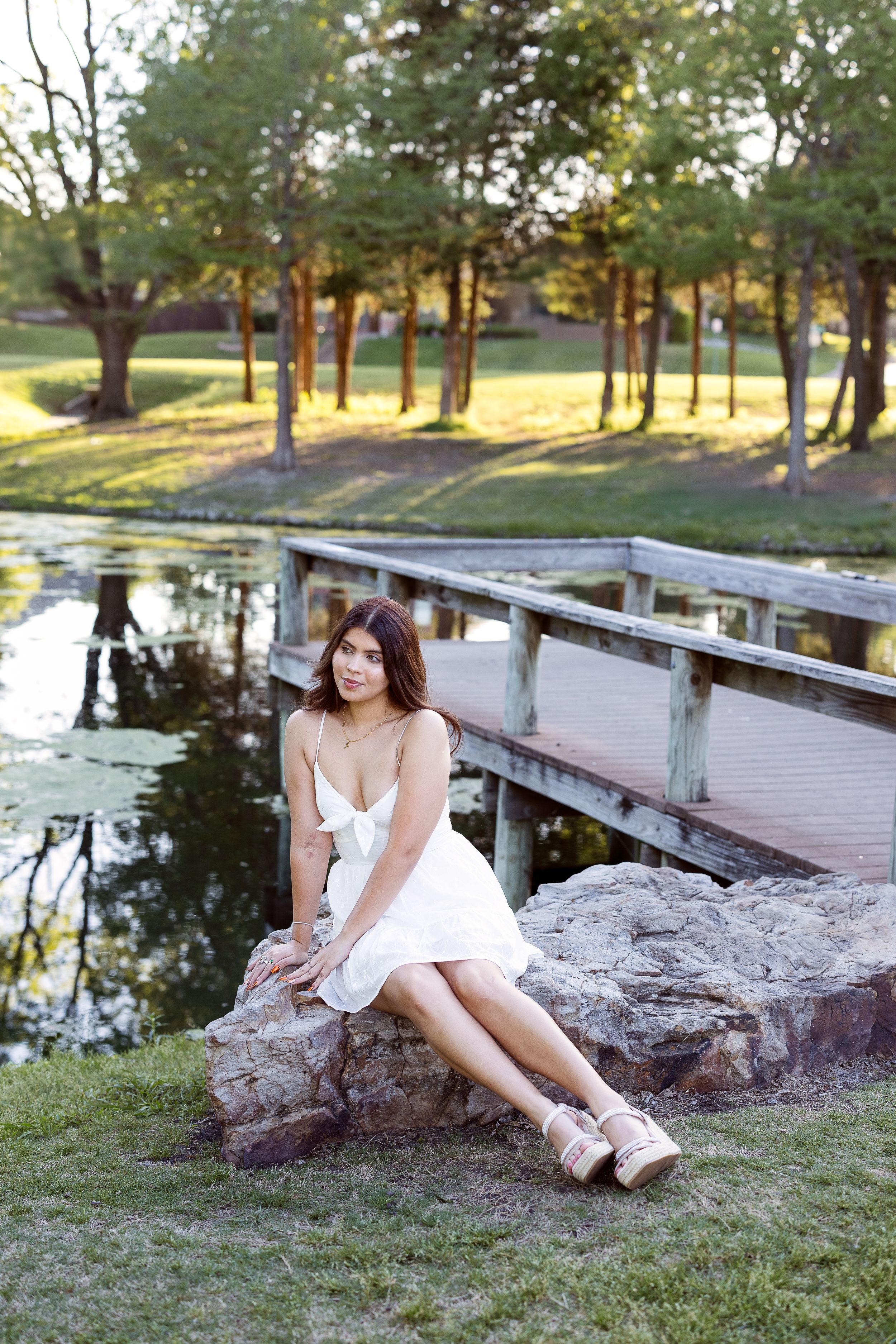 Person in a white dress sits on a large rock by a pond in a scenic park setting at golden hour.