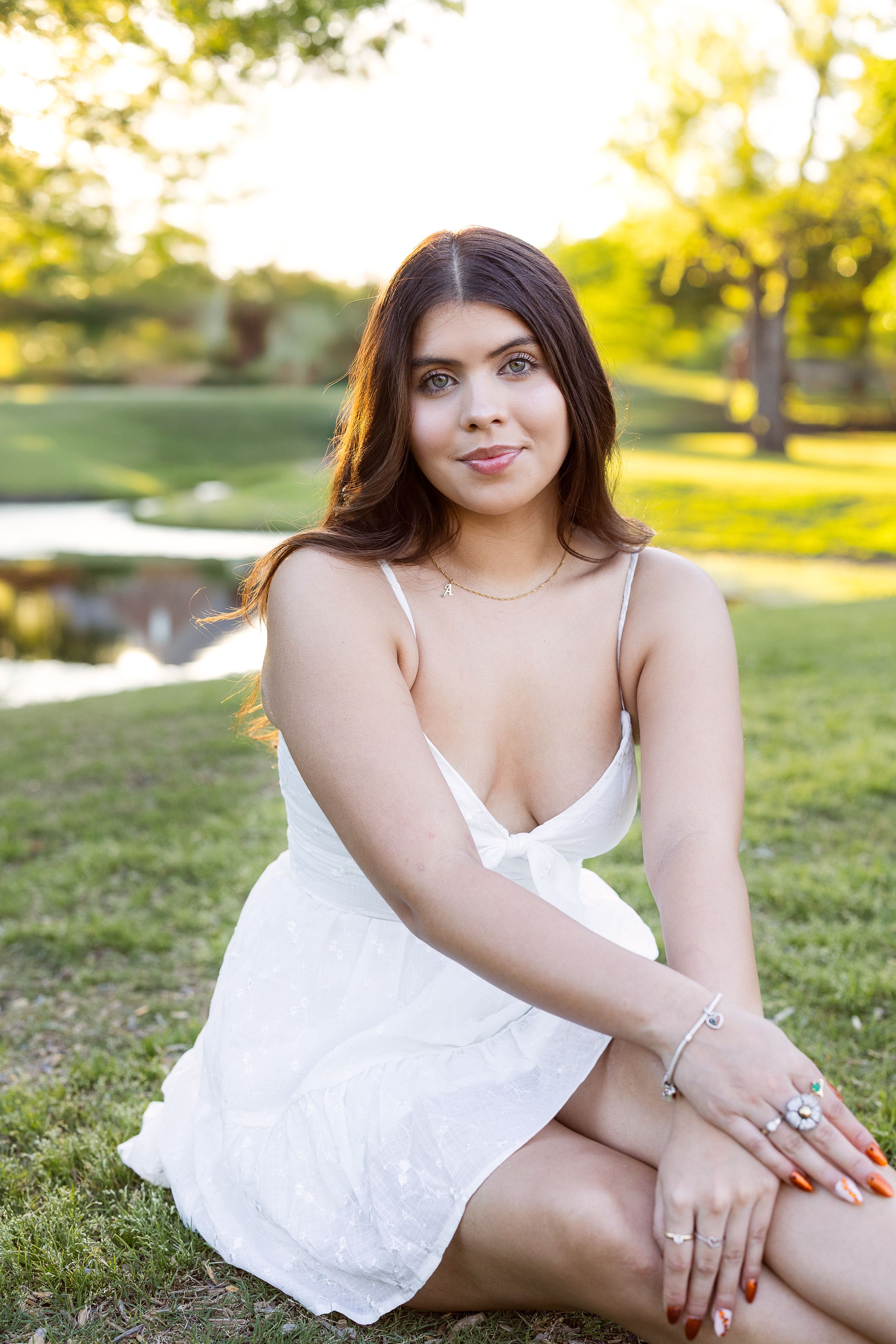 Person in white summer dress sitting on grass in a sunny park with a pond and trees in background.