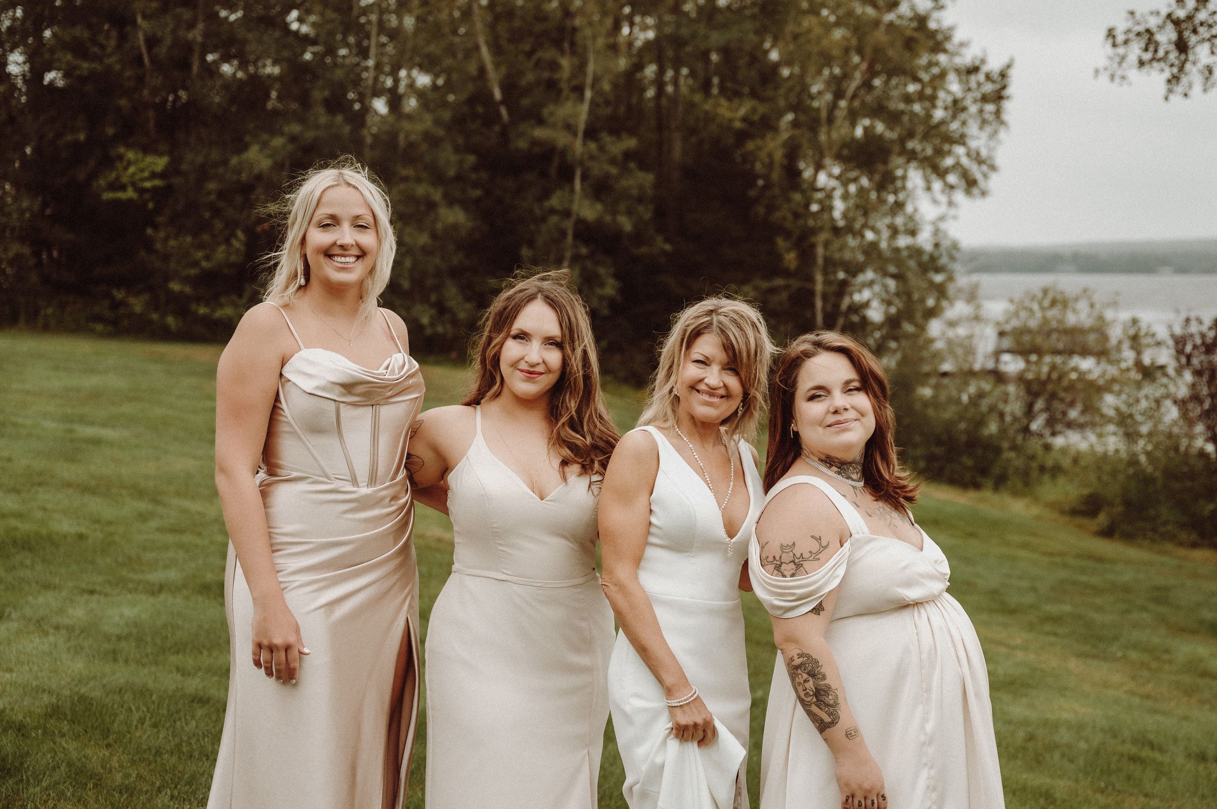 Wedding party in neutral-toned dresses poses together on grassy hillside by lake.