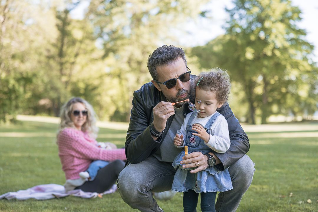 A family shares gentle moments together while sitting on grass in a sunlit park setting.