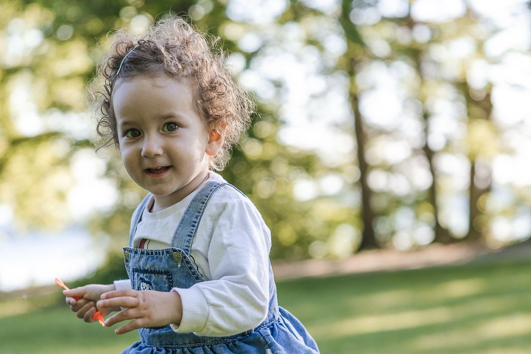 A toddler plays joyfully on grass while wearing a denim outfit and white sweater.