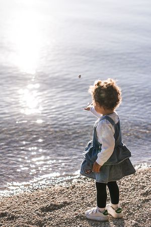 A small child in denim explores a rocky beach shoreline at sunset with water in the background.