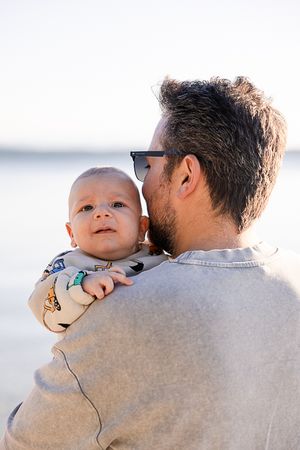 A baby peeking over the shoulder of someone wearing a gray sweater in warm sunlight.