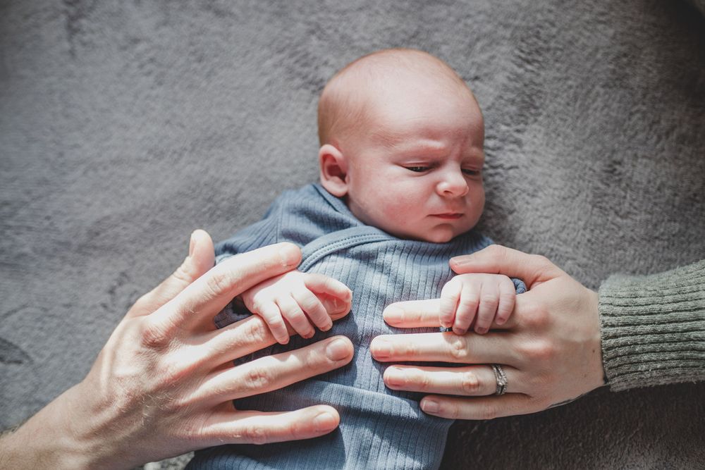 Adult hands gently hold a peaceful baby in a grey sweater against textured grey background.
