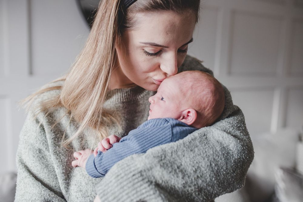 A tender moment between mother and newborn baby as she cradles and kisses the infant in a cozy gray sweater.