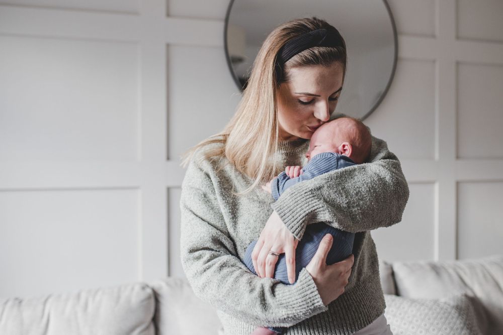 A tender moment captures someone in a cozy gray sweater holding a swaddled newborn baby in a peaceful home setting.