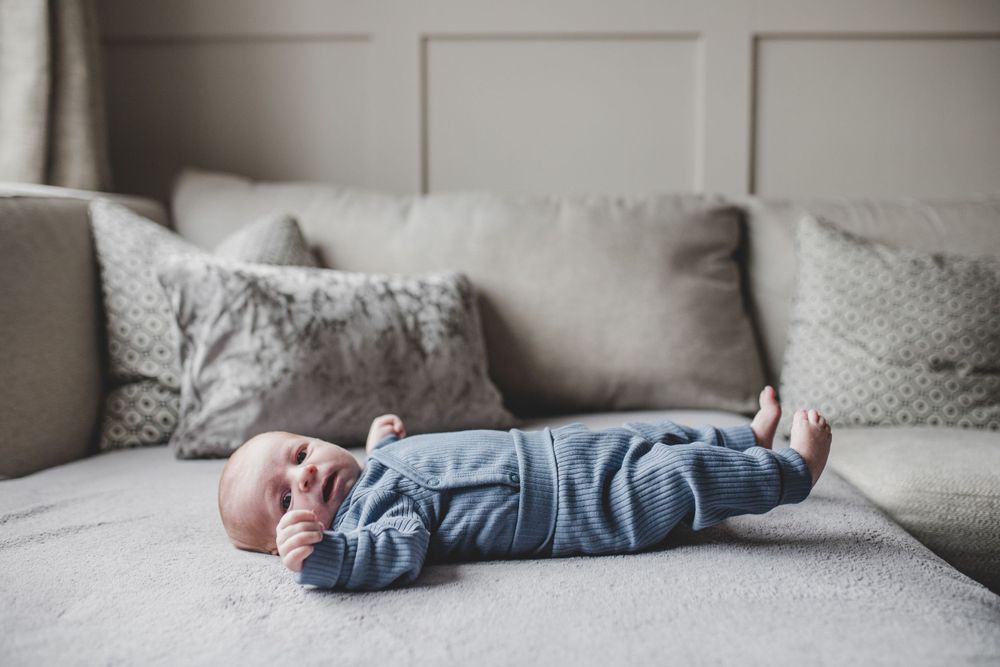 Small baby in denim overalls lies on a gray couch with decorative throw pillows.