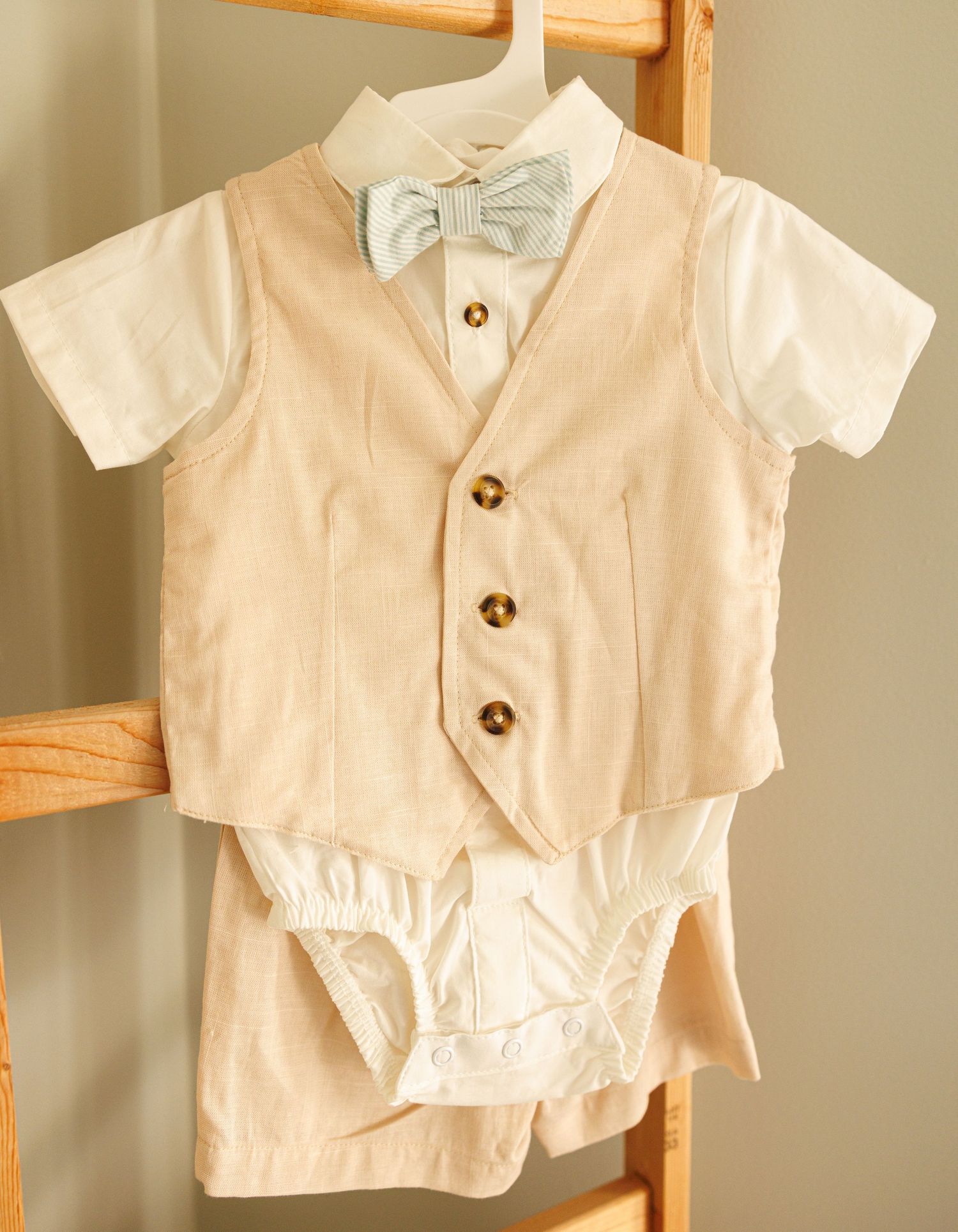 Formal infant outfit with beige vest, white shirt, light blue bow tie, and matching shorts hanging on wooden ladder shelf.