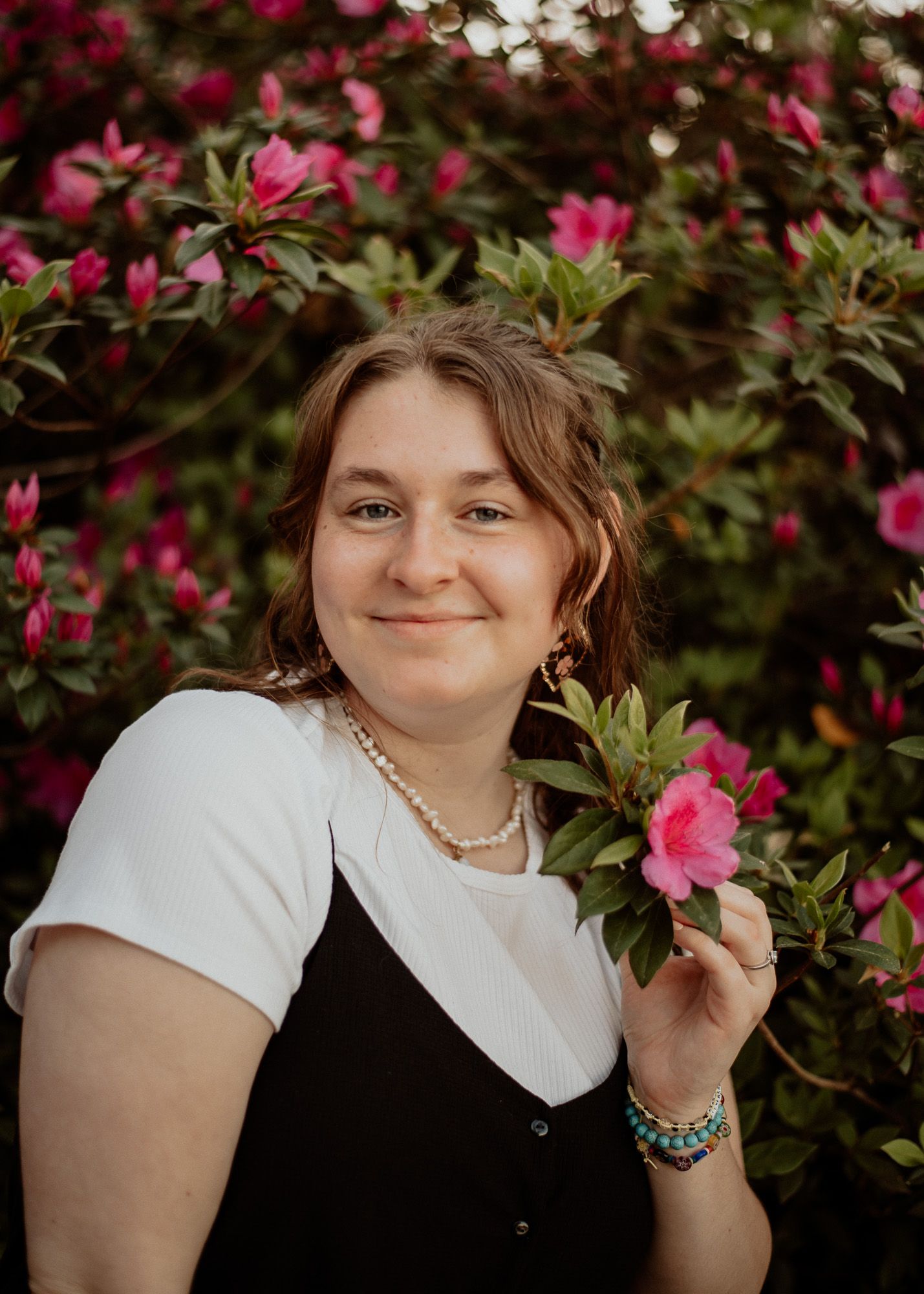 Person poses near pink azalea flowers wearing white shirt and black dress.