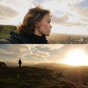 Silhouetted figure stands on a hilltop at sunset with dramatic clouds and cityscape in the distance.