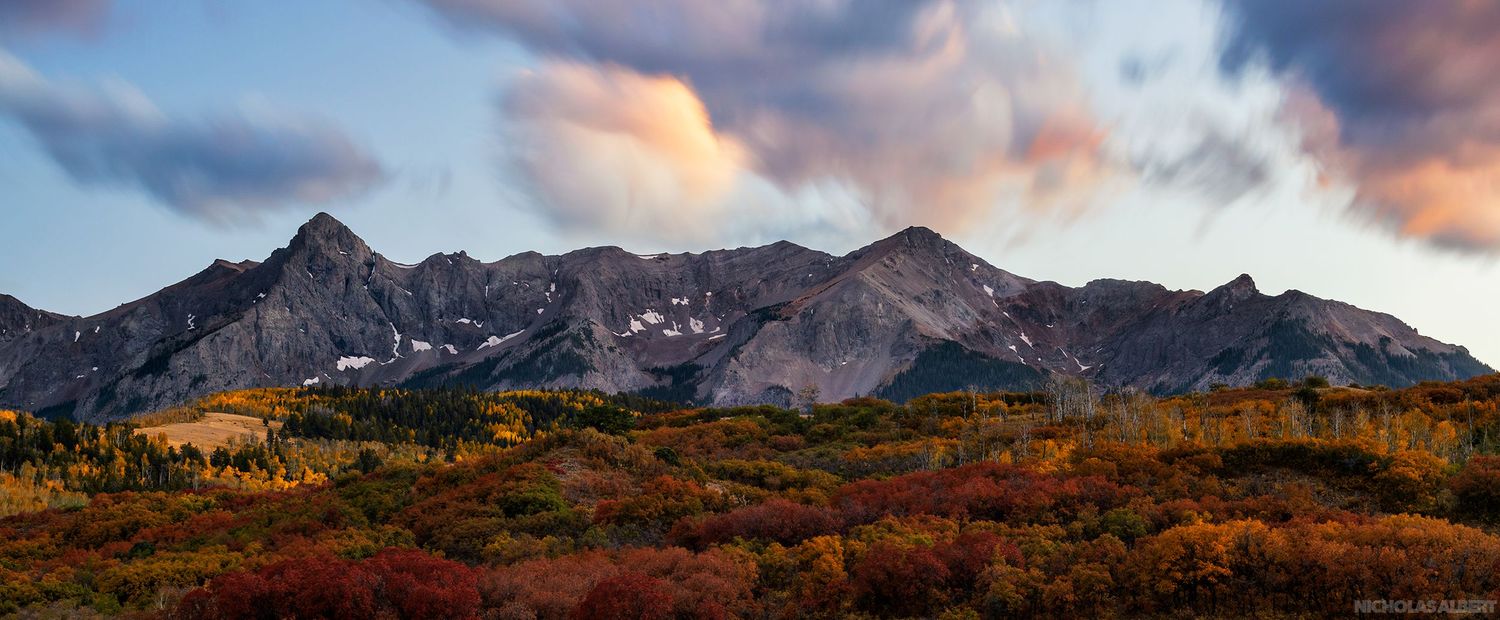 Rocky mountain range with fall colors in foreground under dramatic sunset sky.