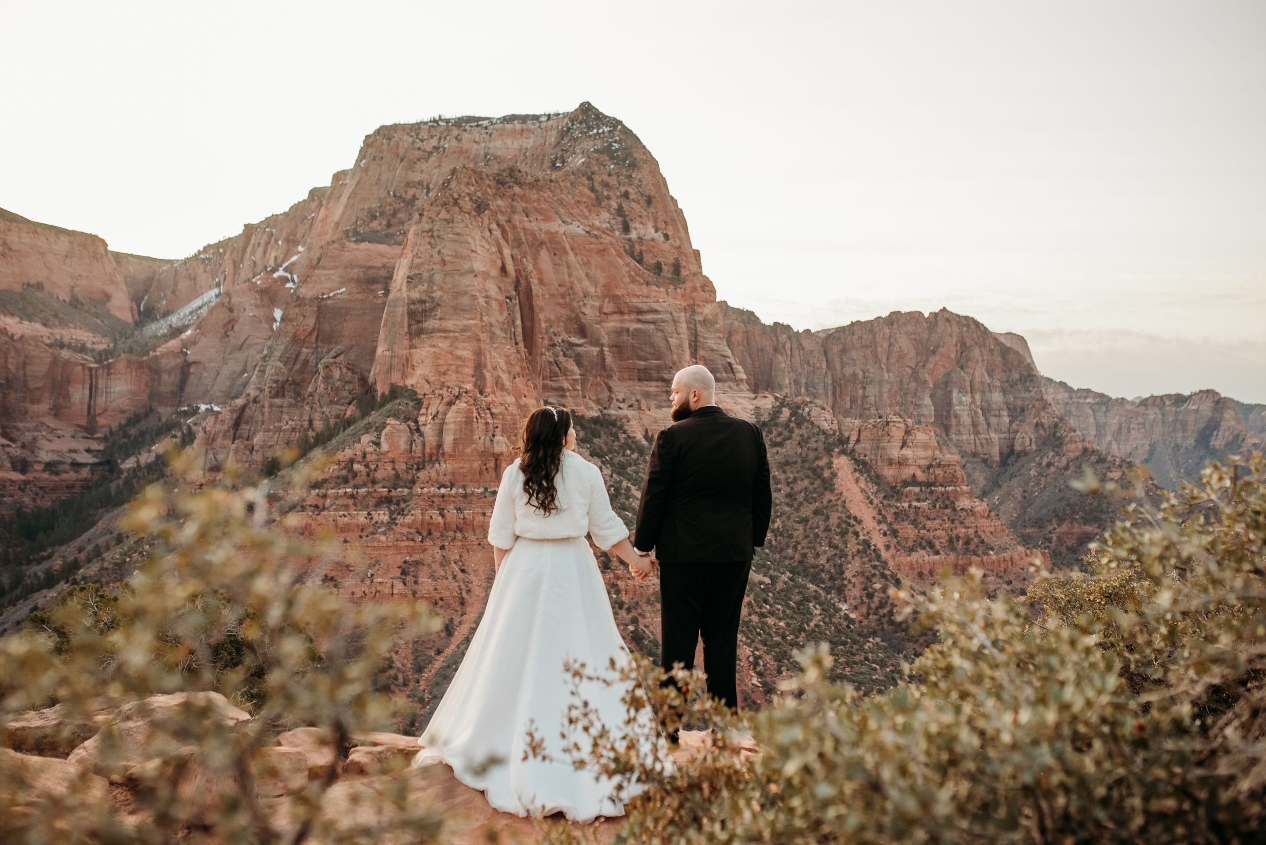 A couple in wedding attire stands together overlooking majestic red rock cliffs at sunset in a national park.