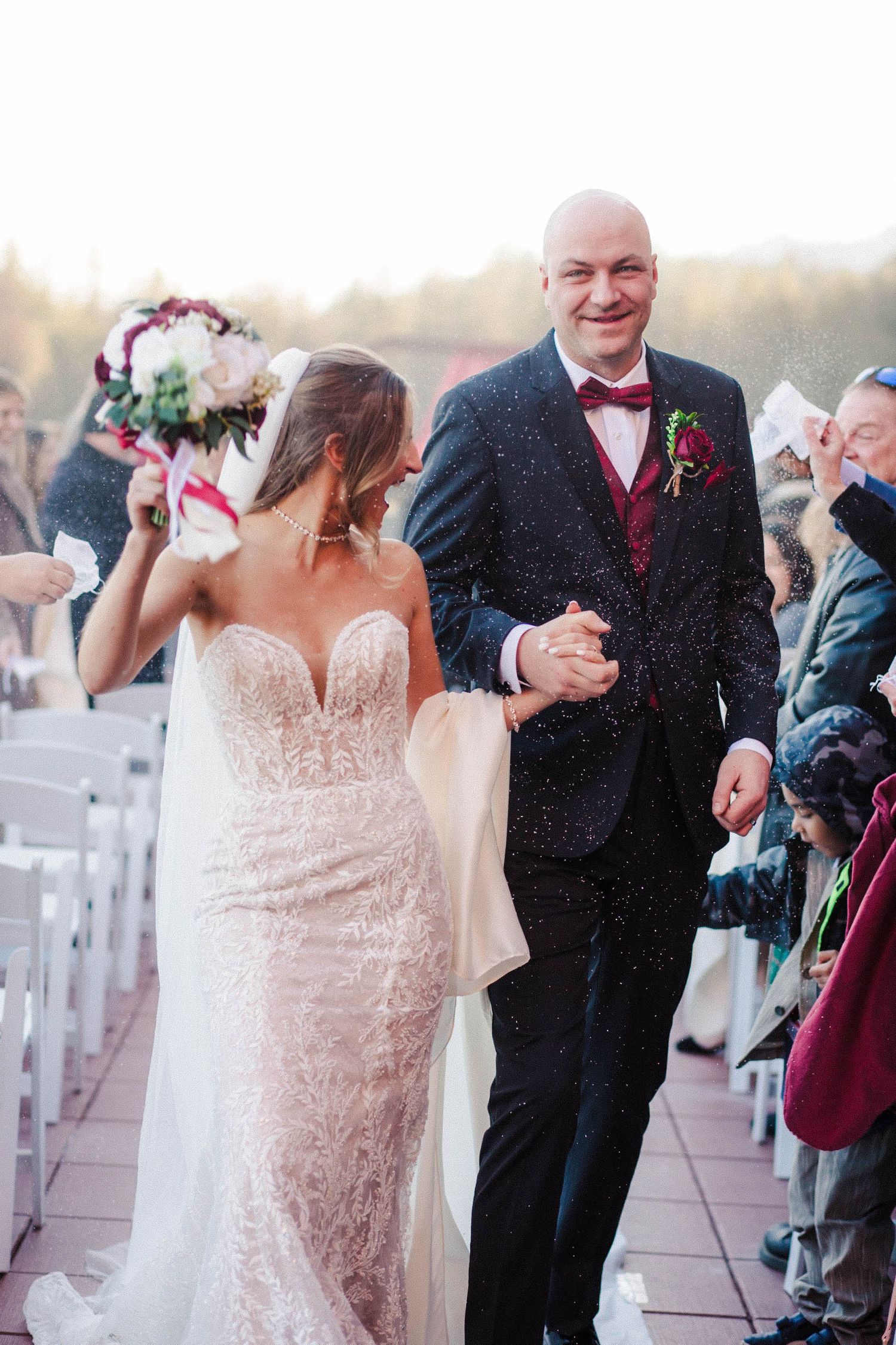 Bride in delicate lace gown walks down aisle with companion in black suit and burgundy bow tie at outdoor ceremony.