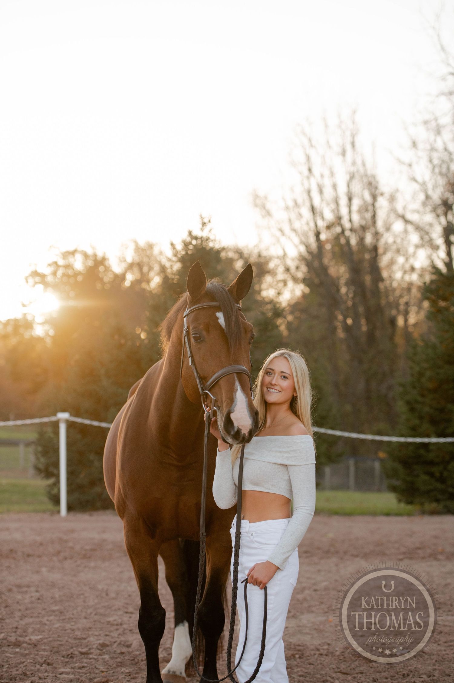 A person in white outfit stands with a bay horse against sunset lighting.