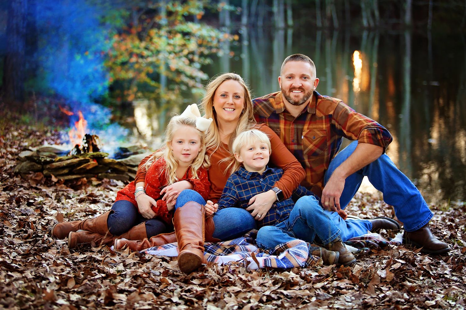A family wearing coordinated outfits sits together on fallen leaves in an autumn forest setting at dusk.