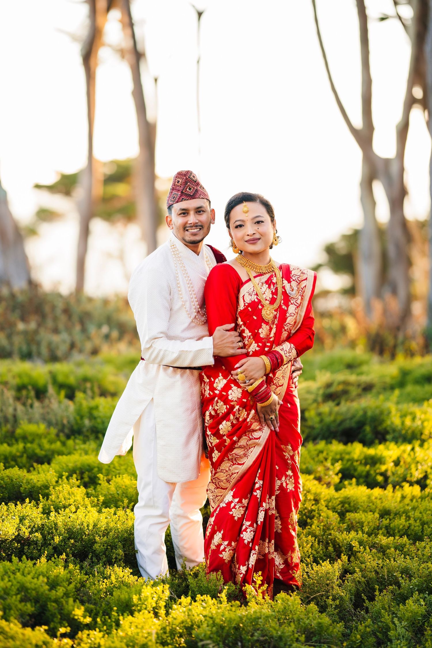 A couple in traditional Nepali wedding attire embraces in a field of yellow wildflowers at sunset.
