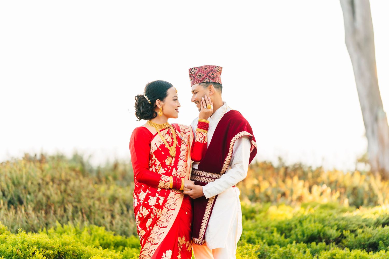 A couple in traditional Nepali wedding attire shares a romantic moment in a sunlit forest.