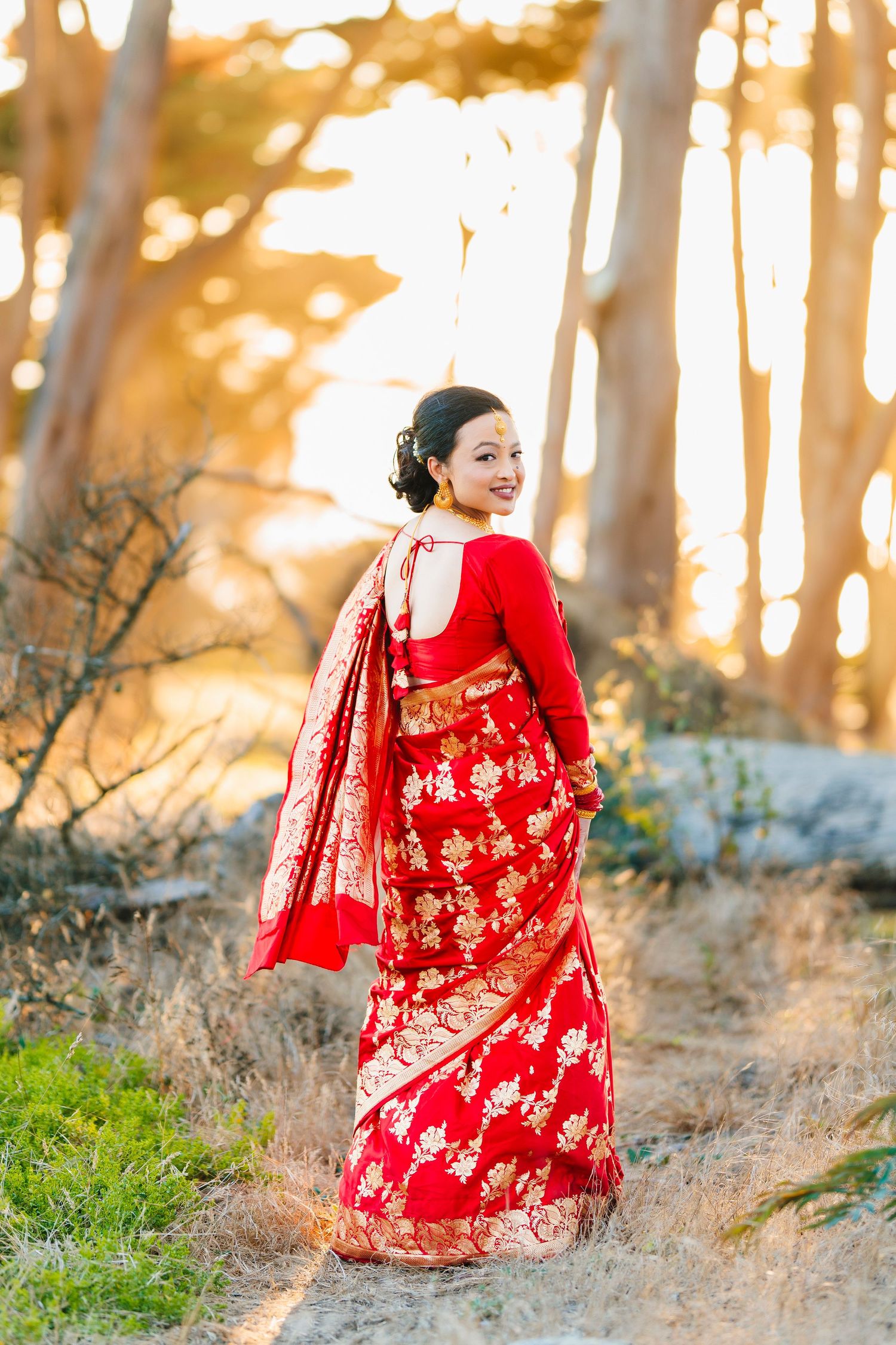 A woman in a red and gold traditional Nepali saree poses in a forest during golden hour.