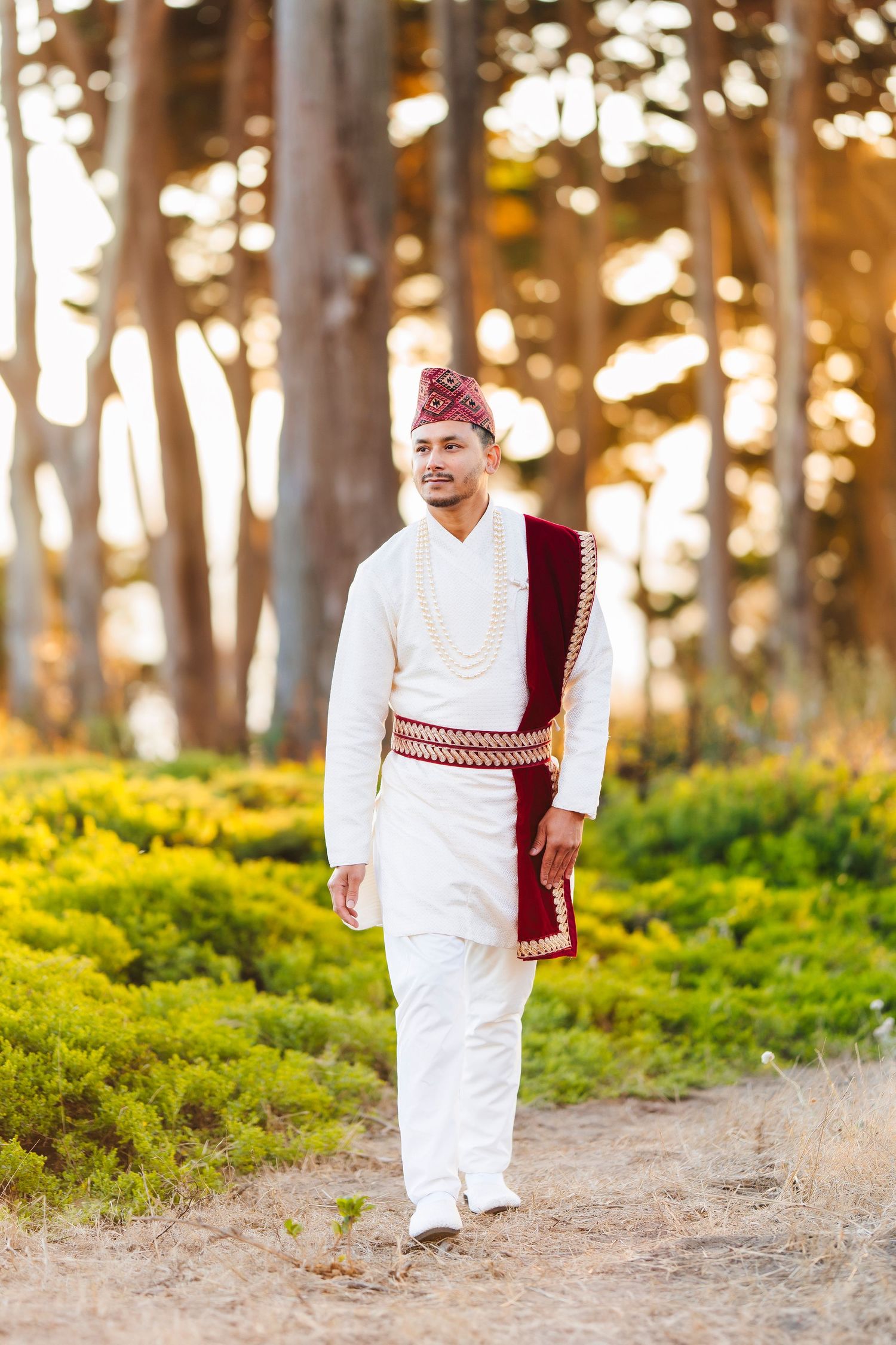 A man in traditional white Nepali wedding attire with burgundy accents walks through a sunlit forest.