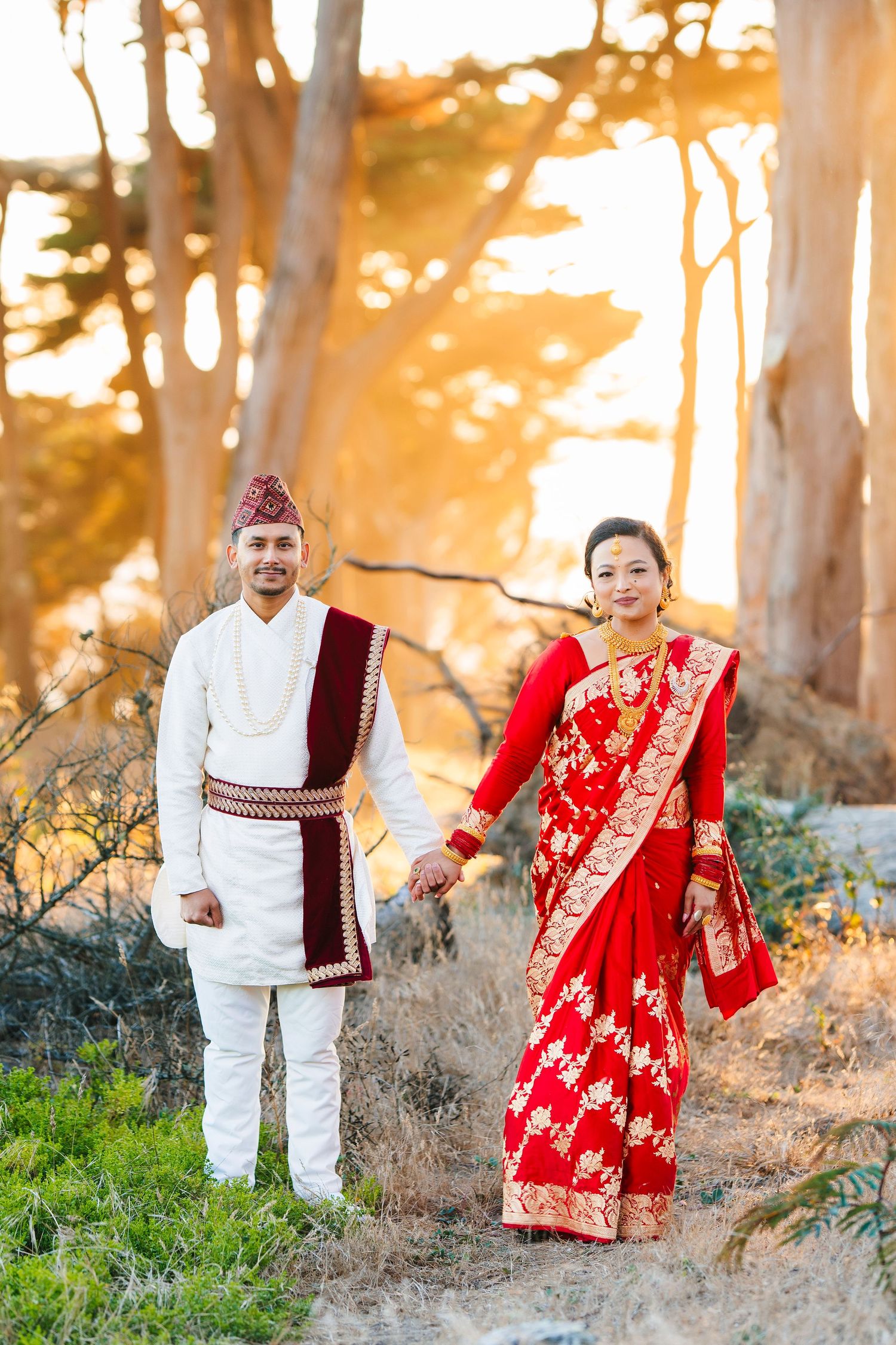 A couple in traditional Nepali wedding attire holds hands while standing in golden evening light.