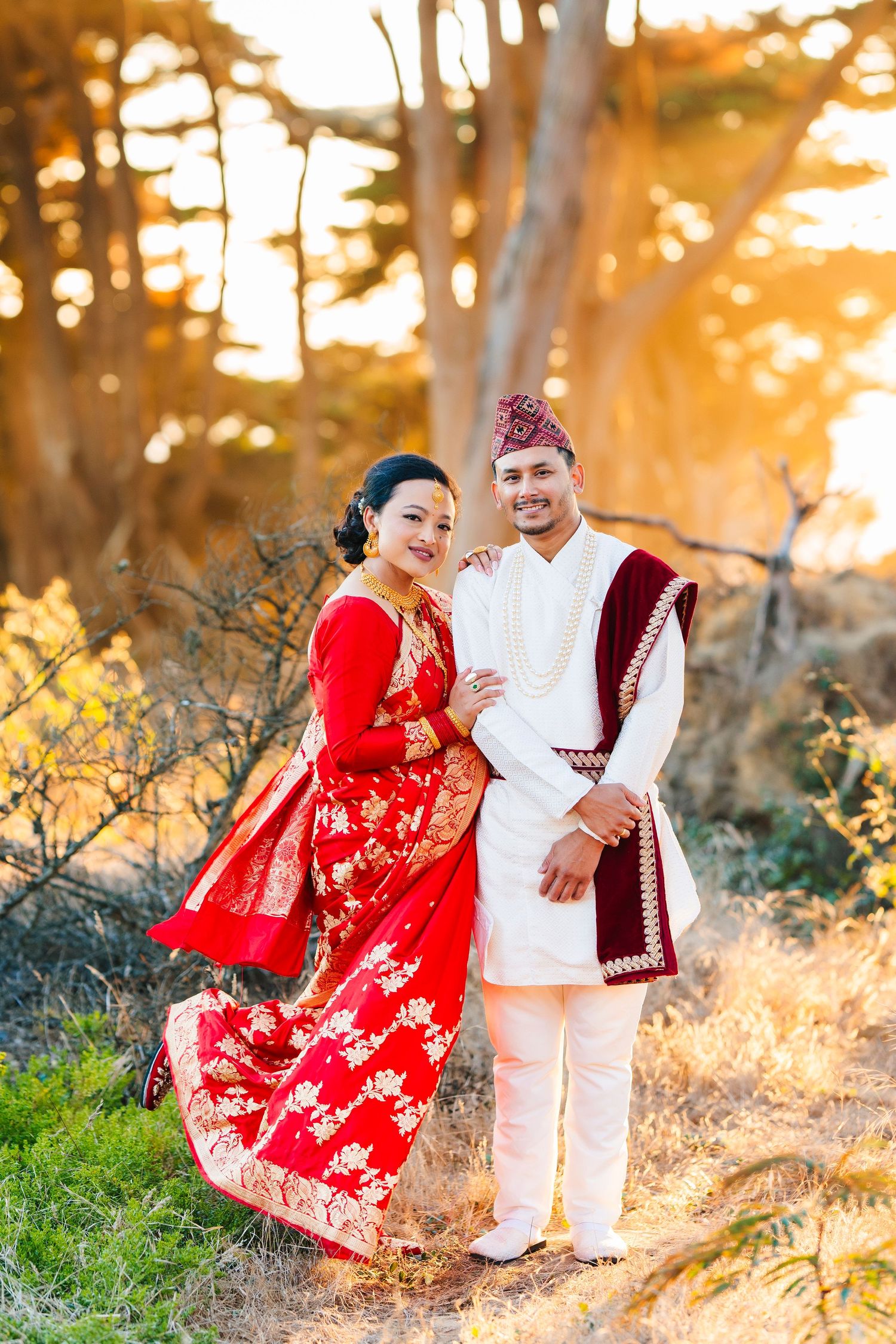 A couple in traditional Nepali wedding clothes poses together amid trees at sunset.