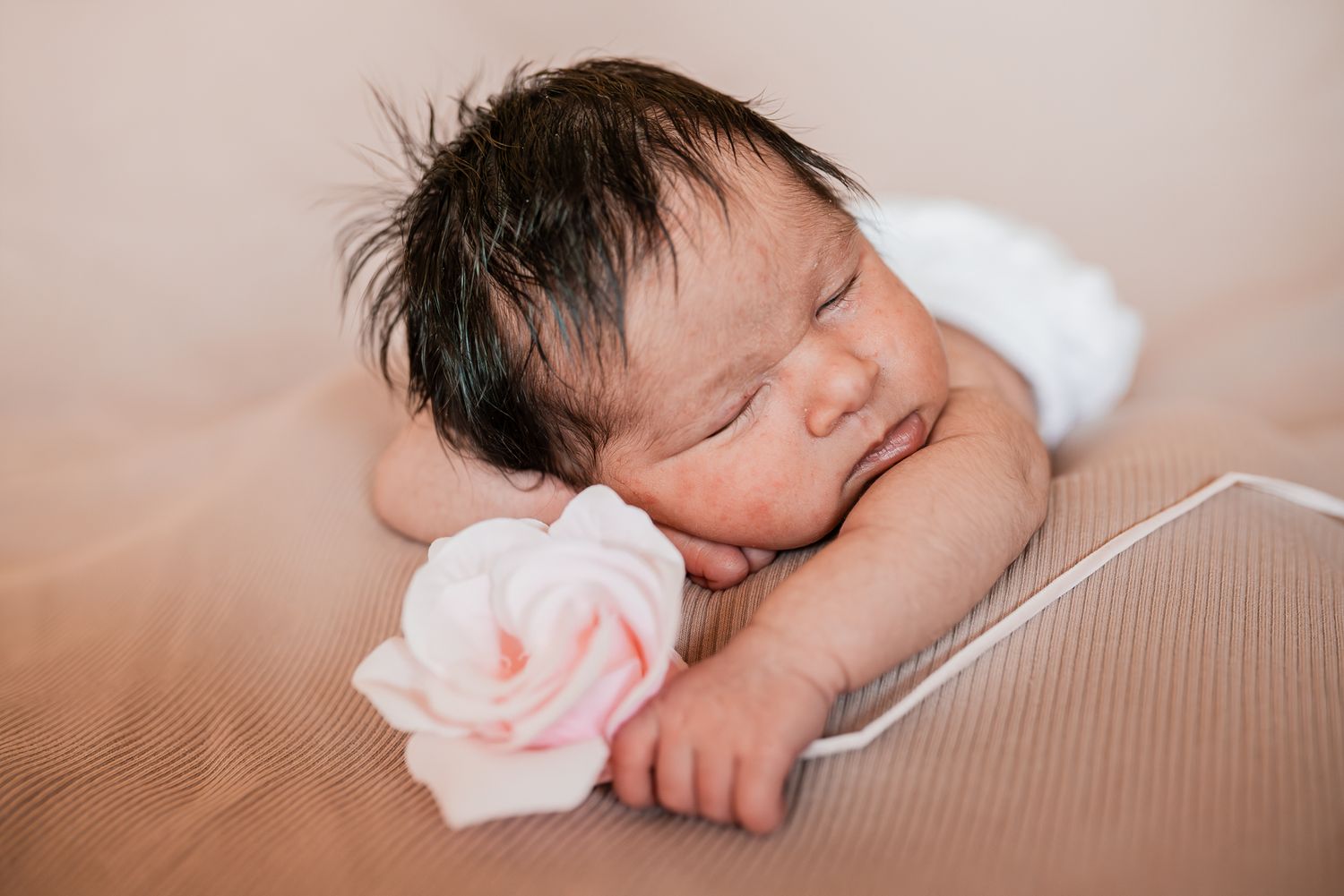 A photo of sleeping newborn baby lies next to a soft pink rose on a beige textured surface  taken by Melbourne newborn photographer.