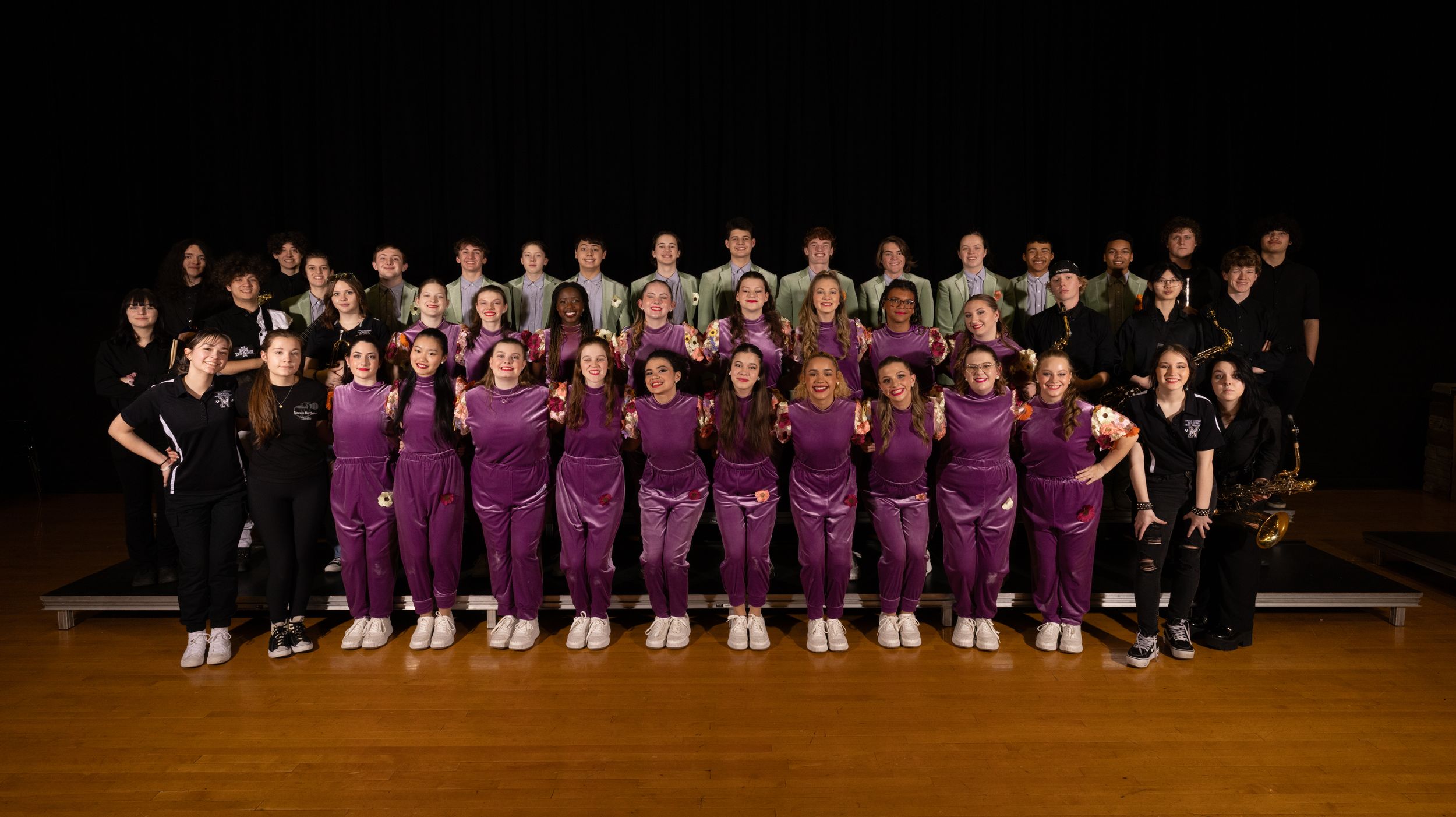 Large dance group in purple costumes performs on wooden stage.