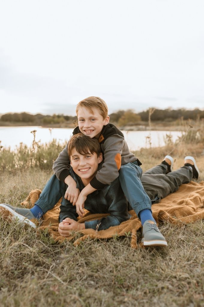 Two people playfully lounging on a brown blanket in a grassy field near a lake on an autumn day.