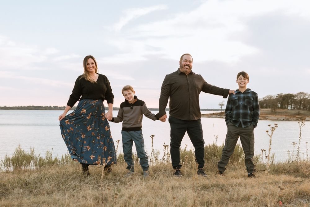 A family poses playfully while sitting on grass near a waterfront during sunset.