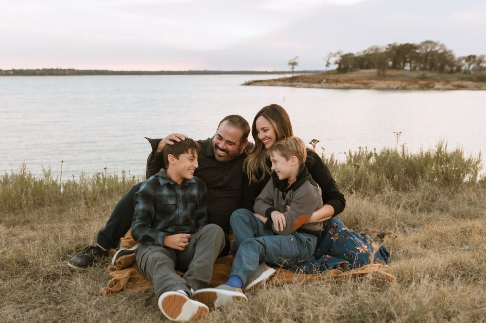 A family relaxes together on grassy terrain near a waterfront during a casual outdoor photoshoot.