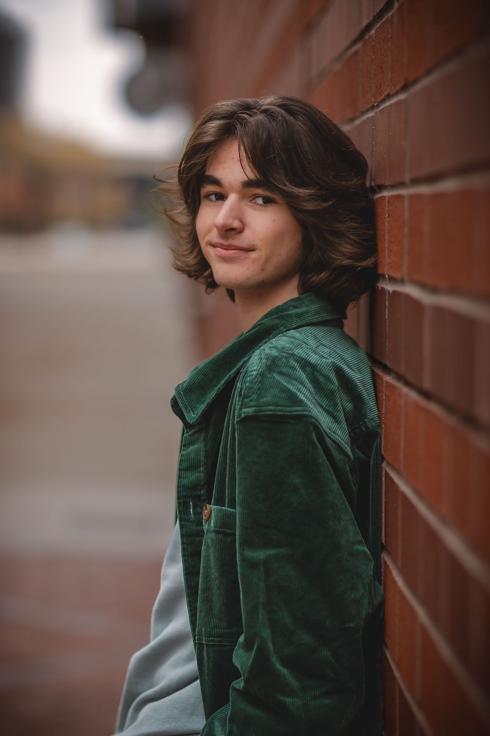 A person in a green corduroy jacket leans against a brick wall while looking away from the camera.