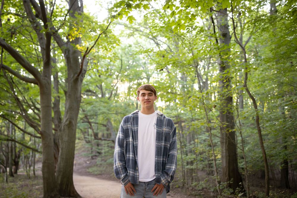 Person in plaid shirt and white tee standing in lush green forest setting.